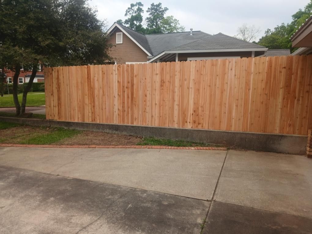 A new, light-colored wooden privacy fence stands atop a low concrete retaining wall in front of a house.