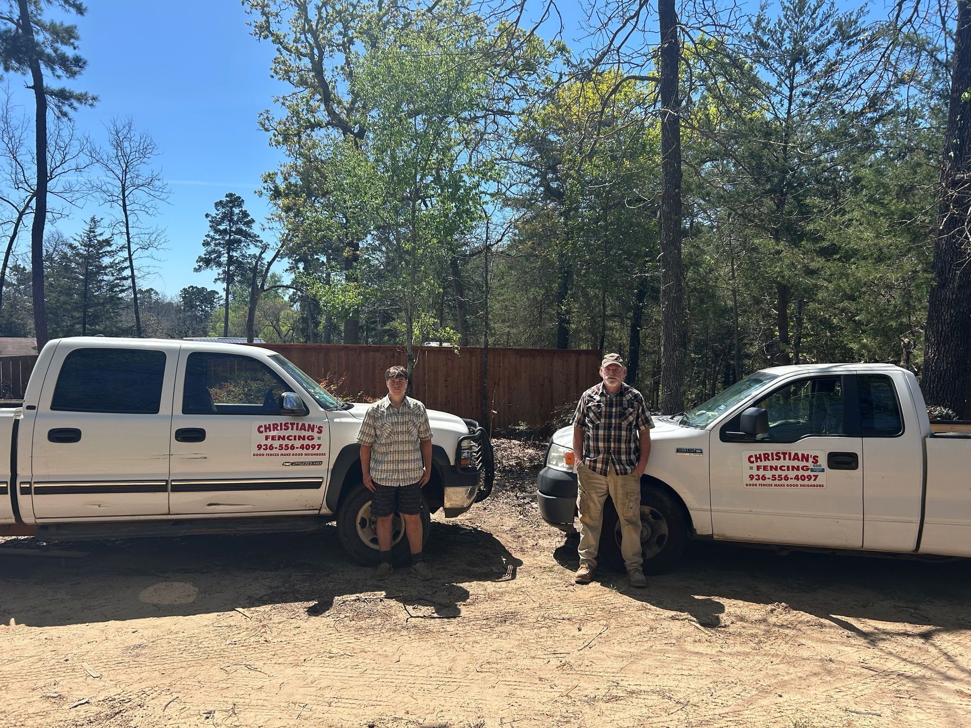Two people stand between two white work trucks parked in a wooded, dirt lot on a sunny day.