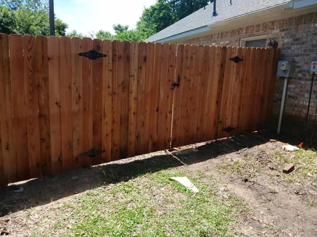 A wooden fence with a double gate and black metal hinges, installed next to a brick house on a sunny day.