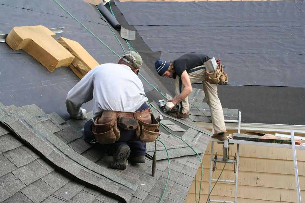 Two roofers installing shingles on a roof. One uses a tool, the other kneels.