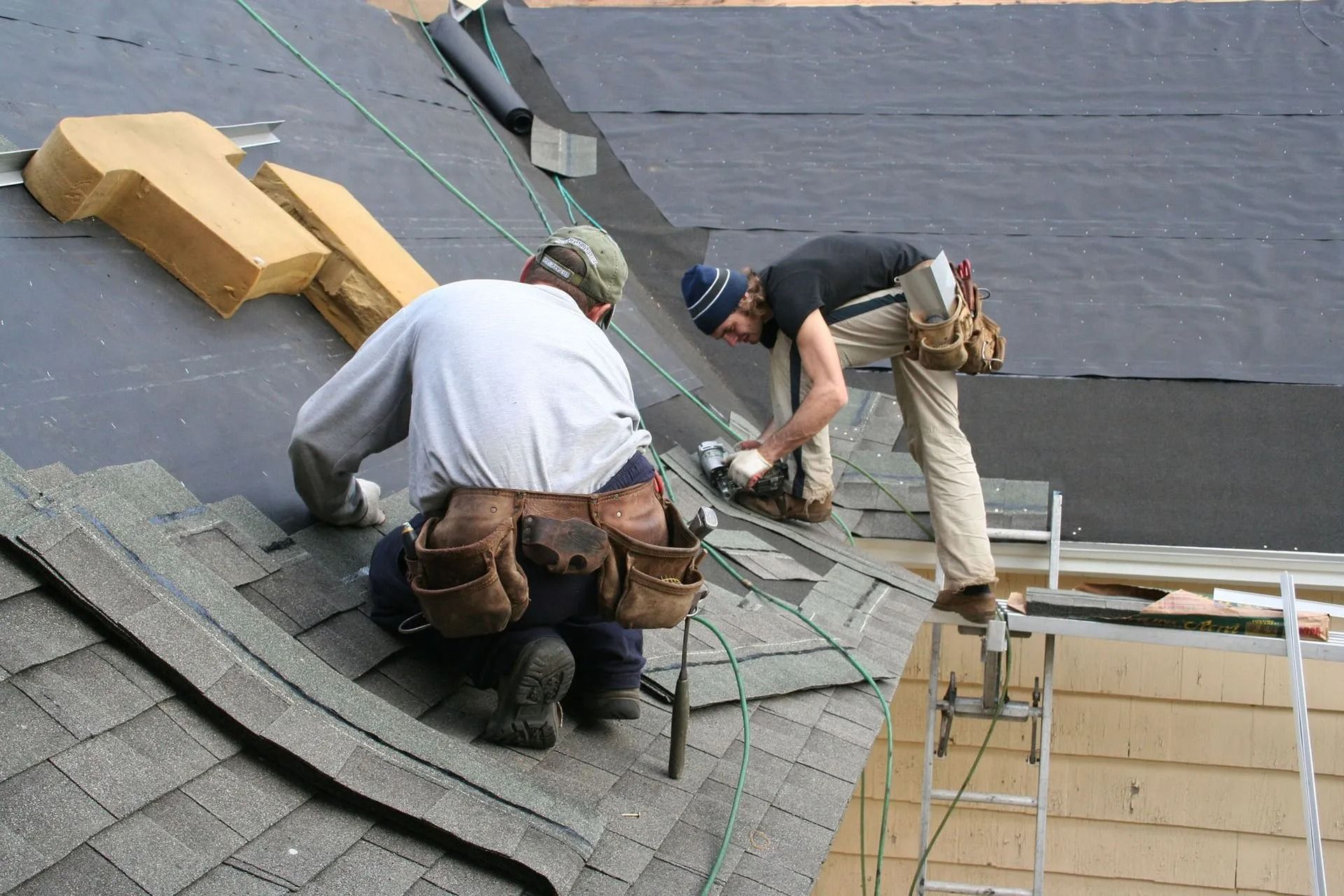 Two roofers installing shingles on a roof. One uses a tool, the other kneels.
