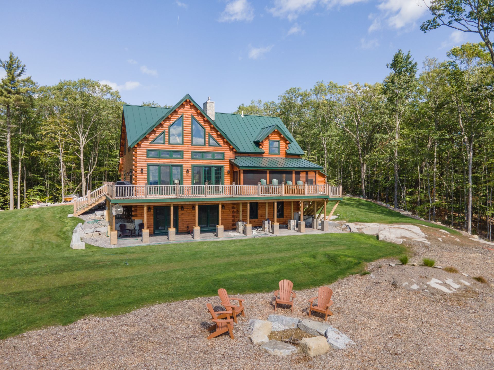 A wooden cabin with a green roof, a large deck, and a stone fire pit with chairs in a grassy clearing surrounded by trees.