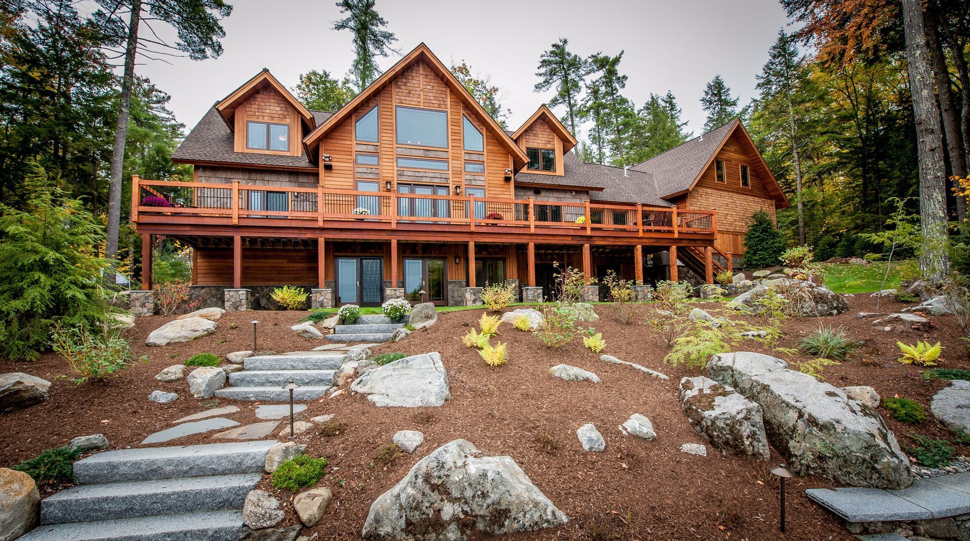 A large, multi-level wooden cabin nestled in a forest, featuring a wide deck, stone steps, and a landscaped yard.