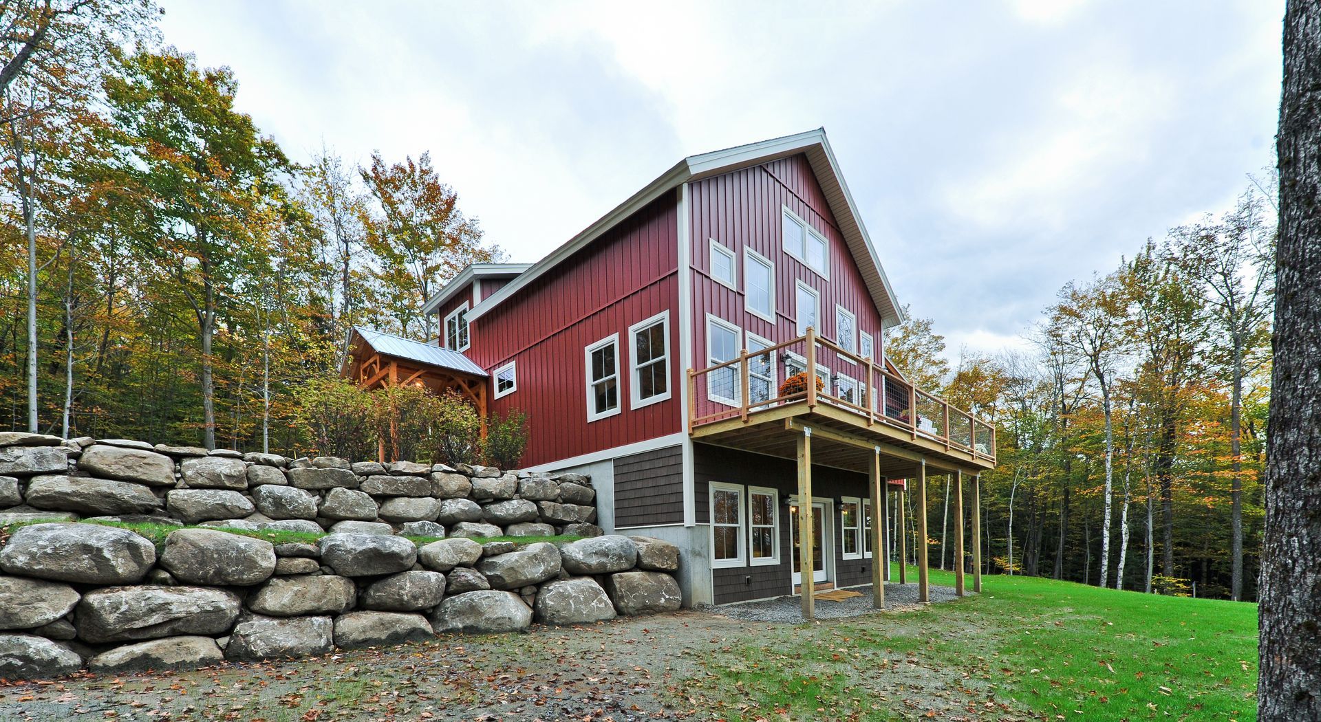Red barn-style house with stone retaining wall, wooden deck, surrounded by trees and a green lawn under an overcast sky.