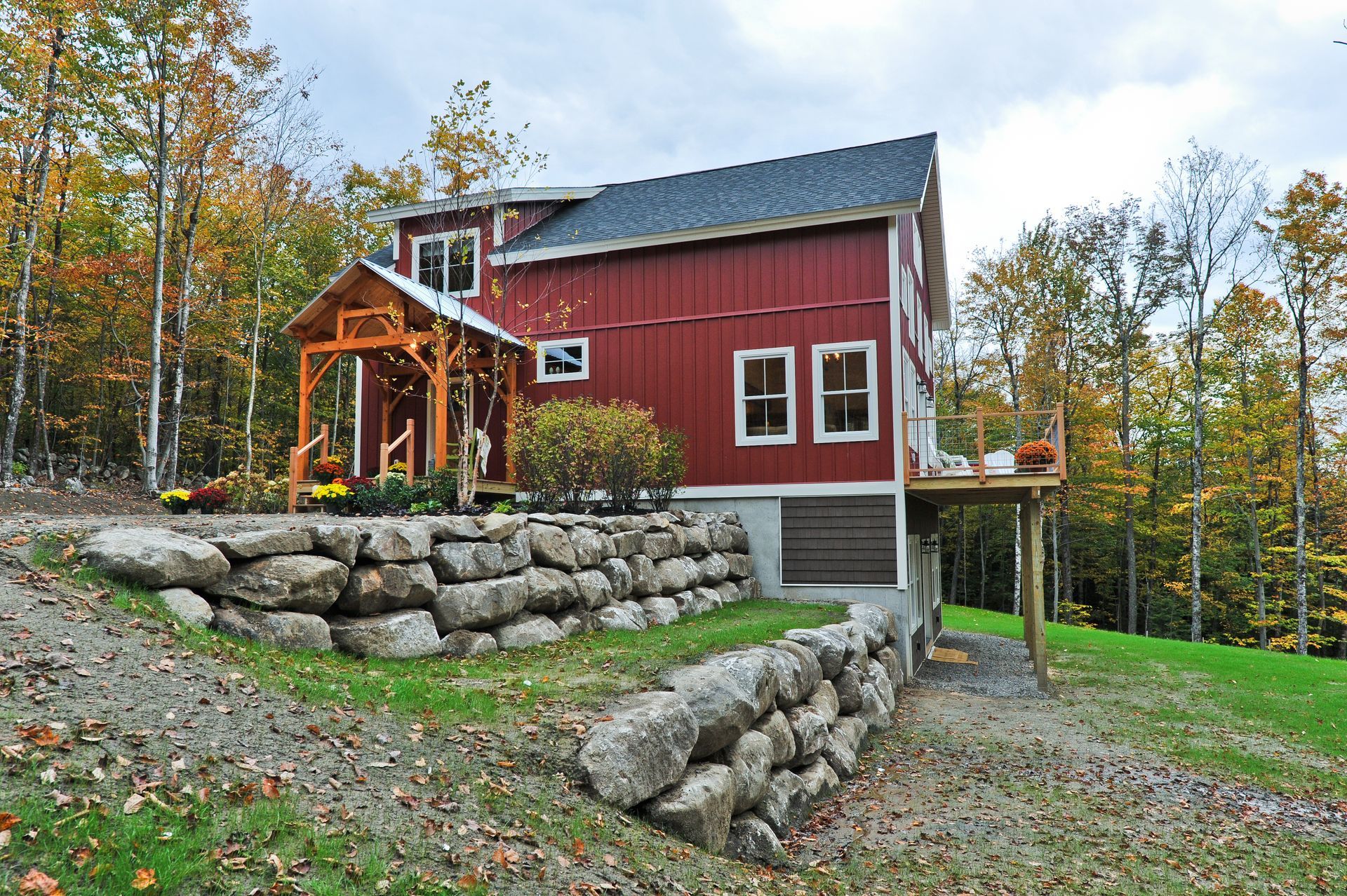 Red, two-story house with a wooden porch, on a hill with a rock retaining wall and autumn trees.