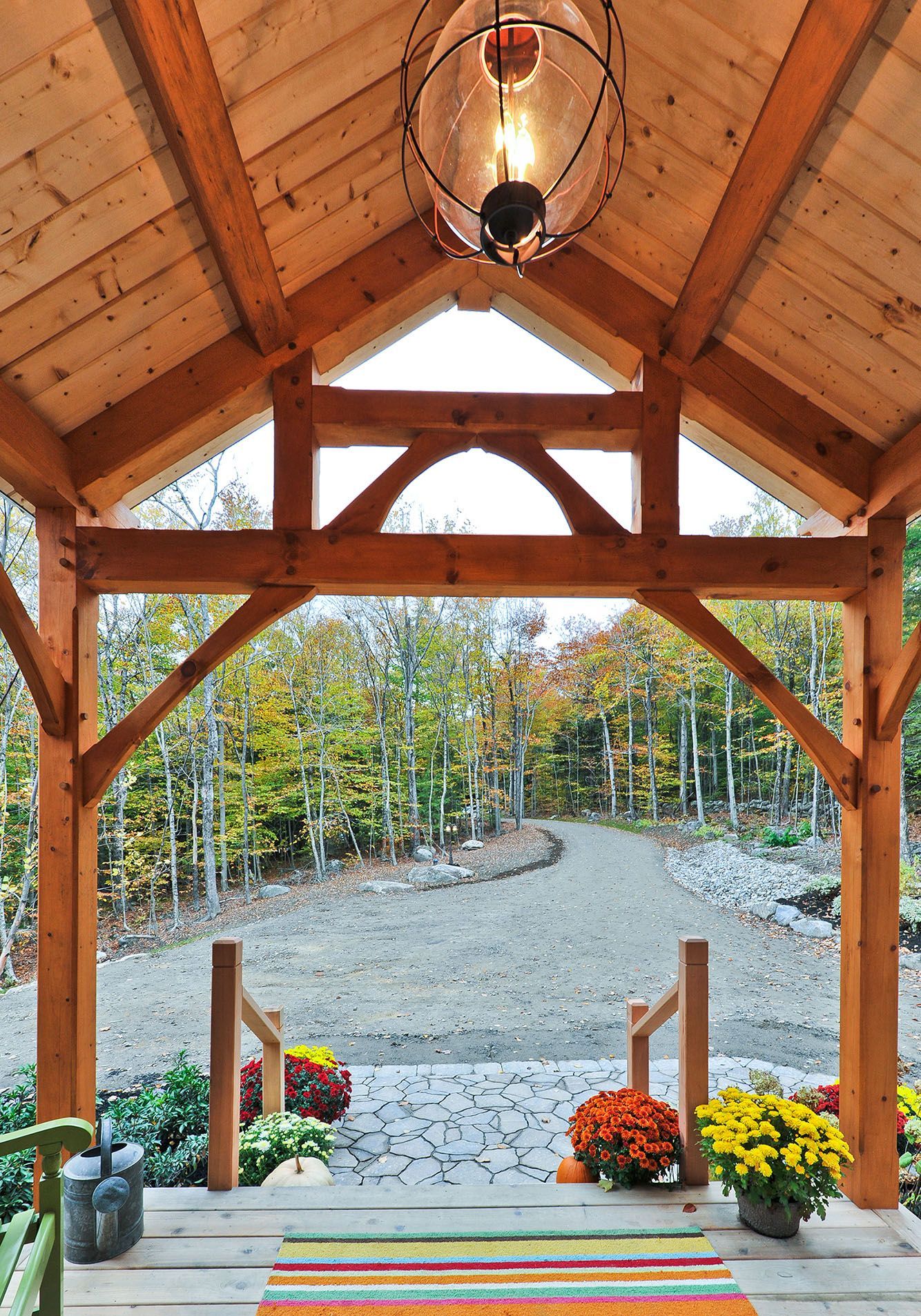 Wooden porch entrance with fall foliage views. Porch has a rug, mums, and a hanging light.