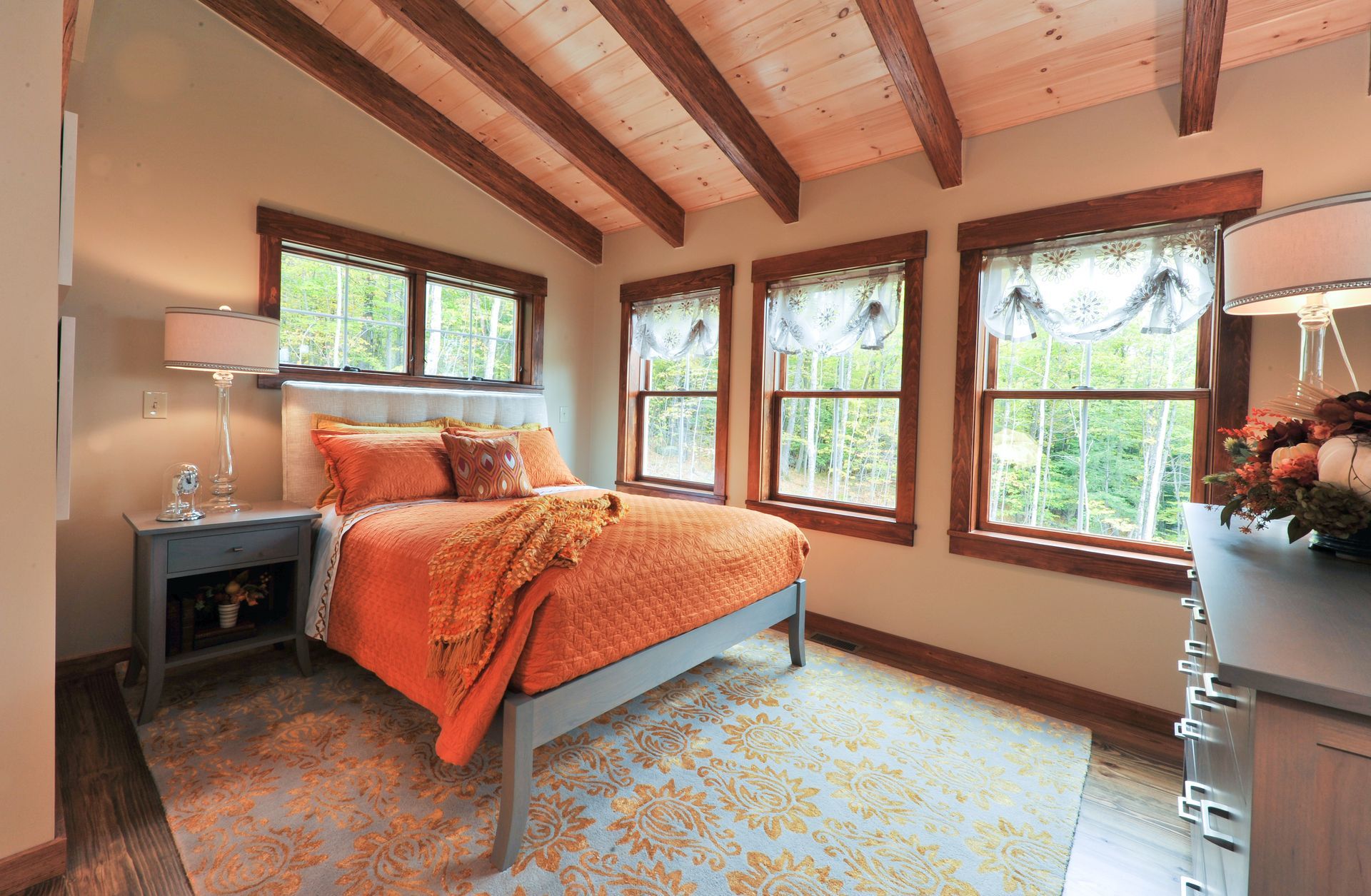 Bedroom with orange bedding, windows, wooden beams, nightstand with lamp, dresser.