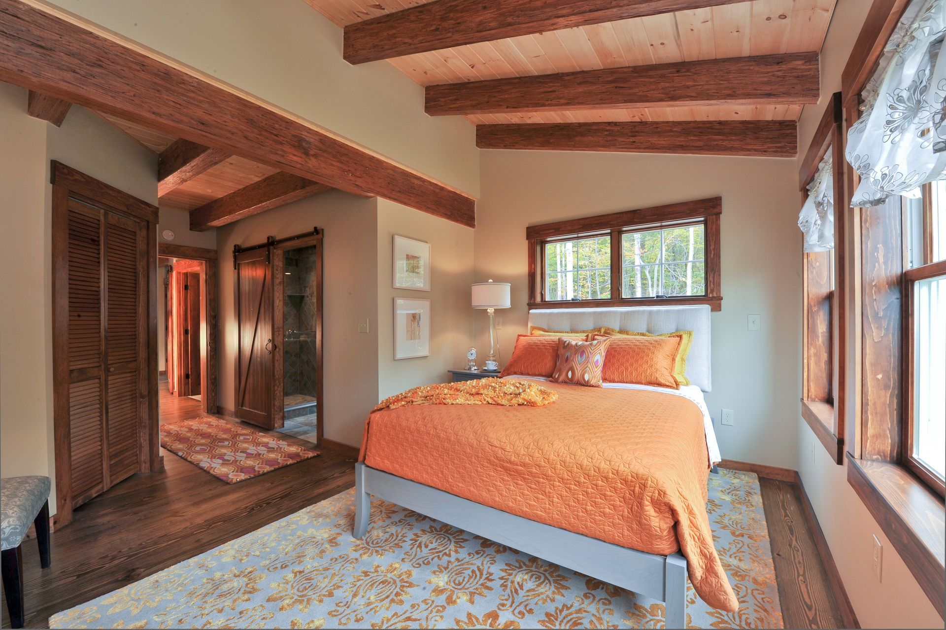 Bedroom with orange bedspread, wood beams, and large windows.
