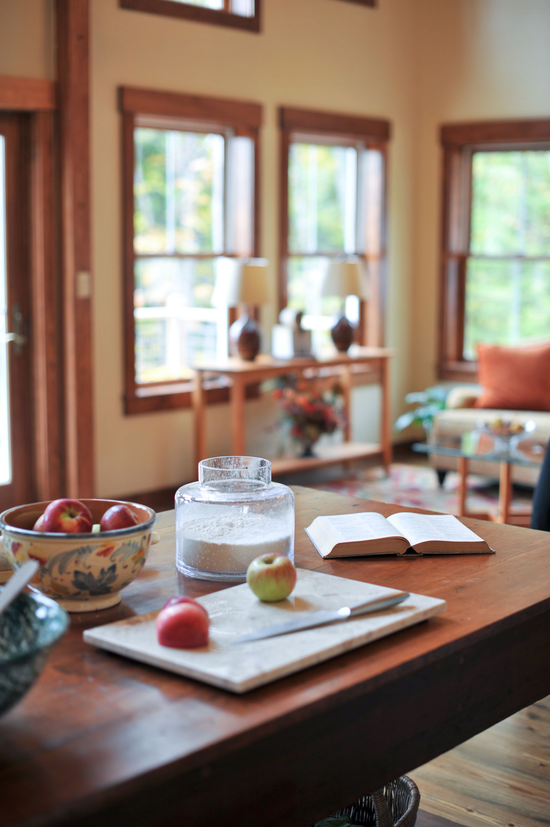 Kitchen with table holding fruit, cutting board, book, and jar, with windows in background.