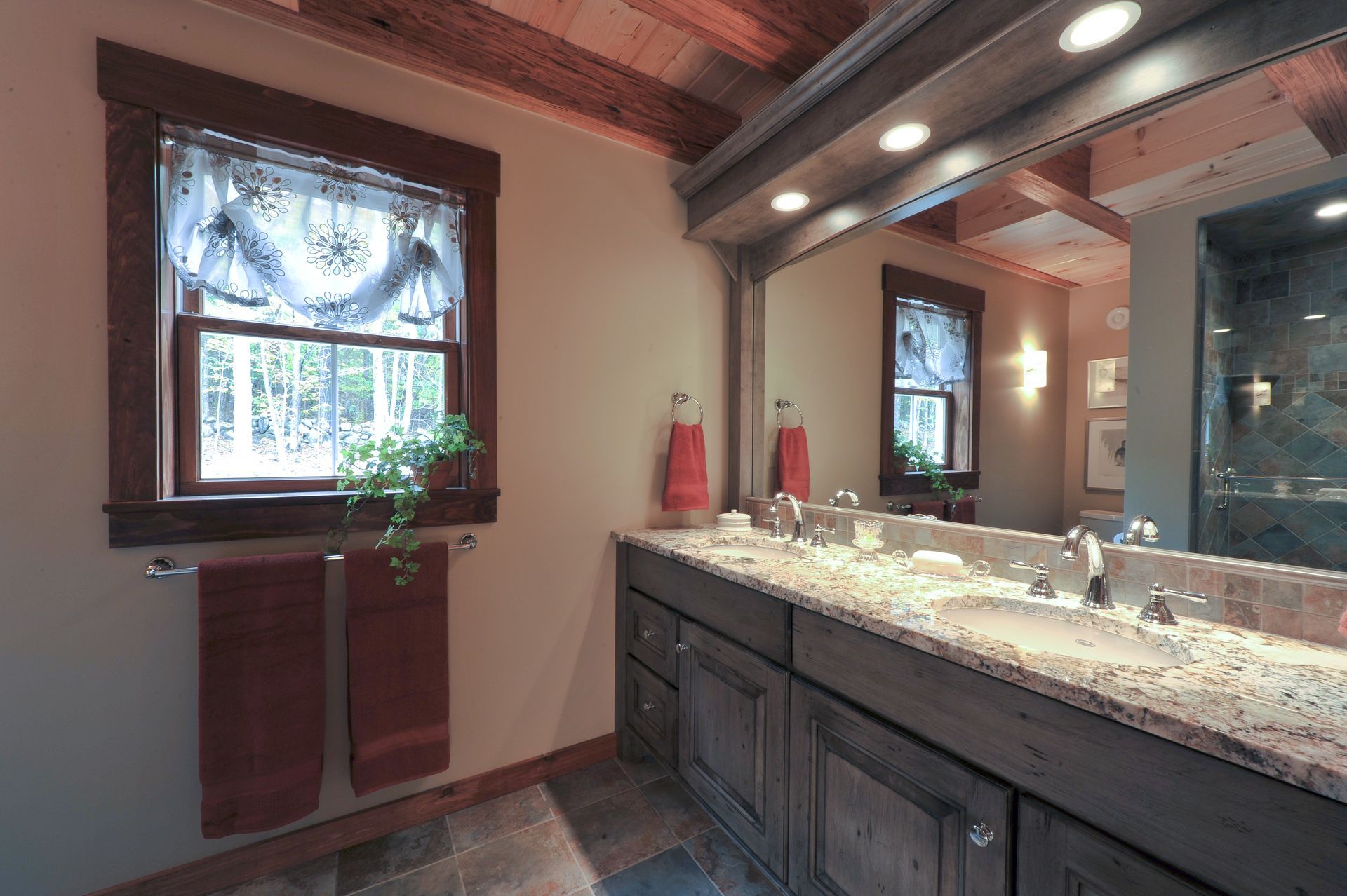 Rustic bathroom with double sink, wooden cabinets, window with curtains, and two orange towels.