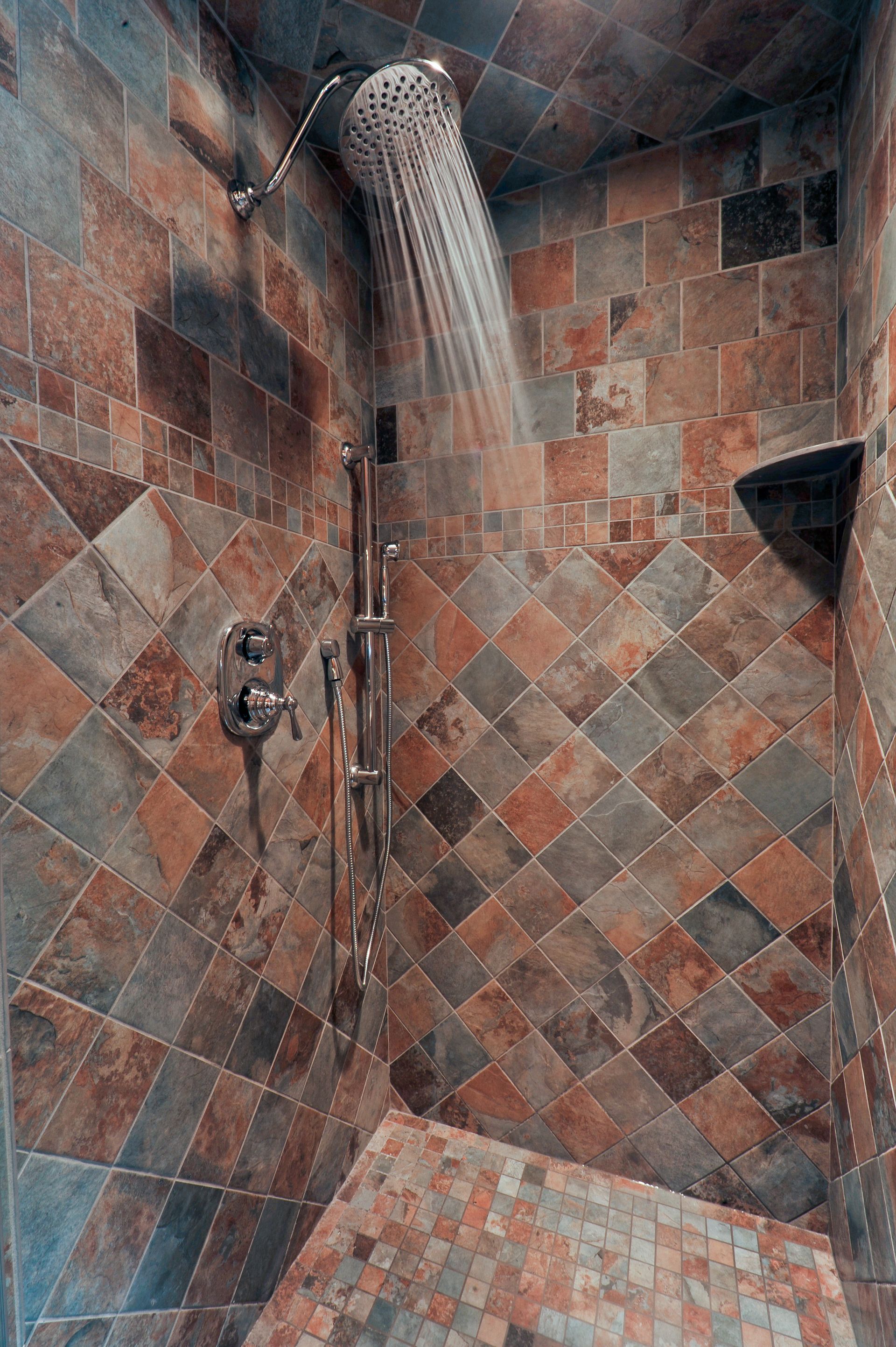 Shower with patterned brown and gray tiles, water flowing from the overhead shower head, small shelf in wall.