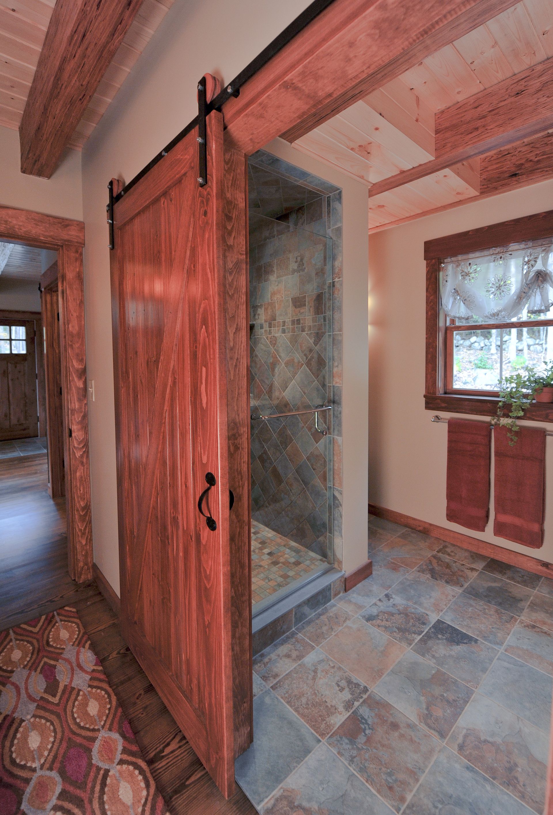 Barn door leads to a shower in a rustic bathroom with stone tile flooring and exposed wooden beams.
