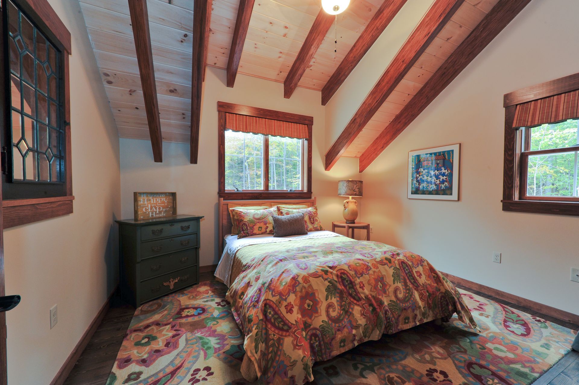 Bedroom with angled ceiling, exposed beams, bed with patterned comforter, and windows.