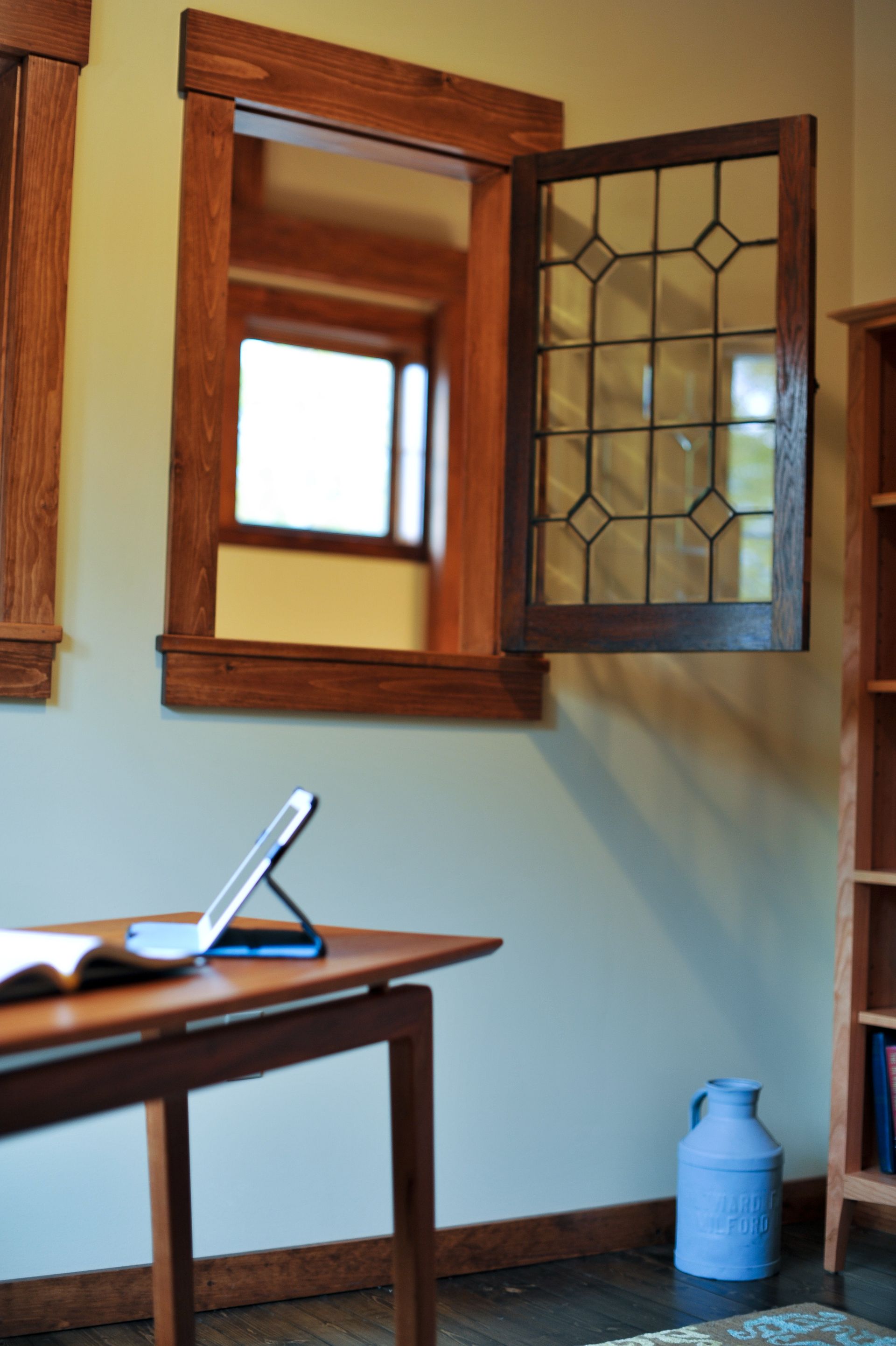 Wooden-framed window with stained glass panes, open next to a desk with a tablet and a blue vase in a room.