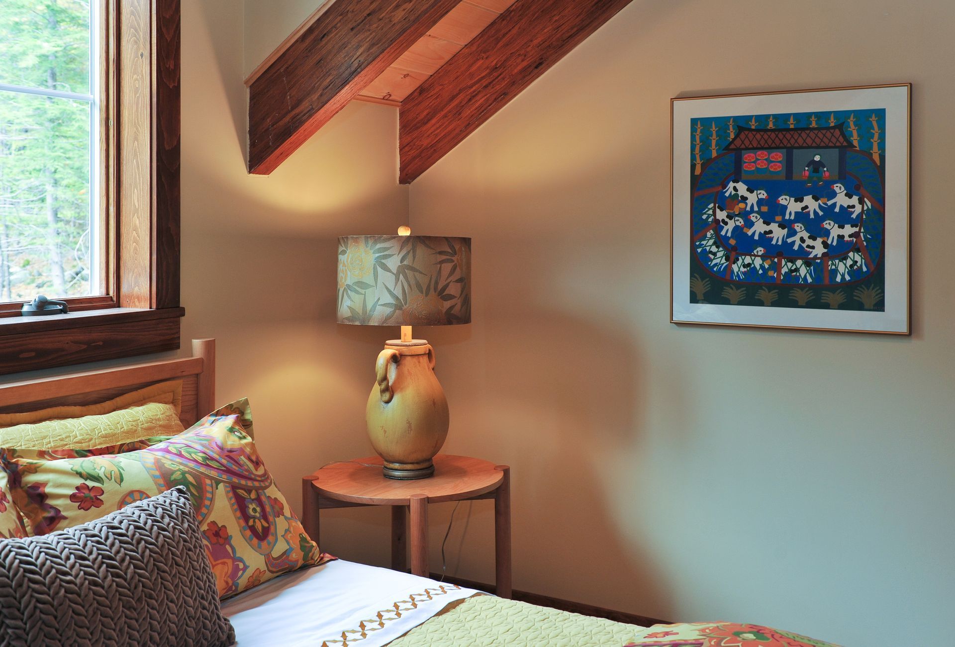 Cozy bedroom with bed, bedside table, lamp, and framed folk art. Window on left. Warm colors, natural light.