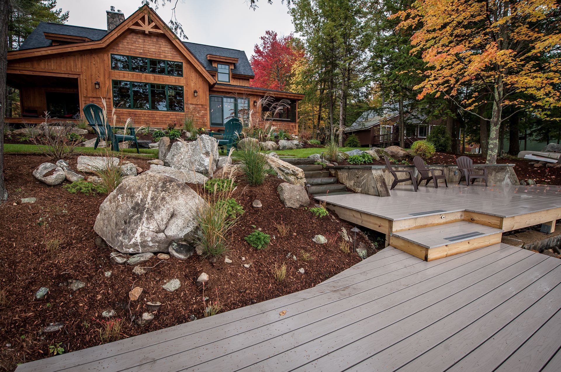 Wooden lakeside home with a composite deck, large boulders, and fall foliage.