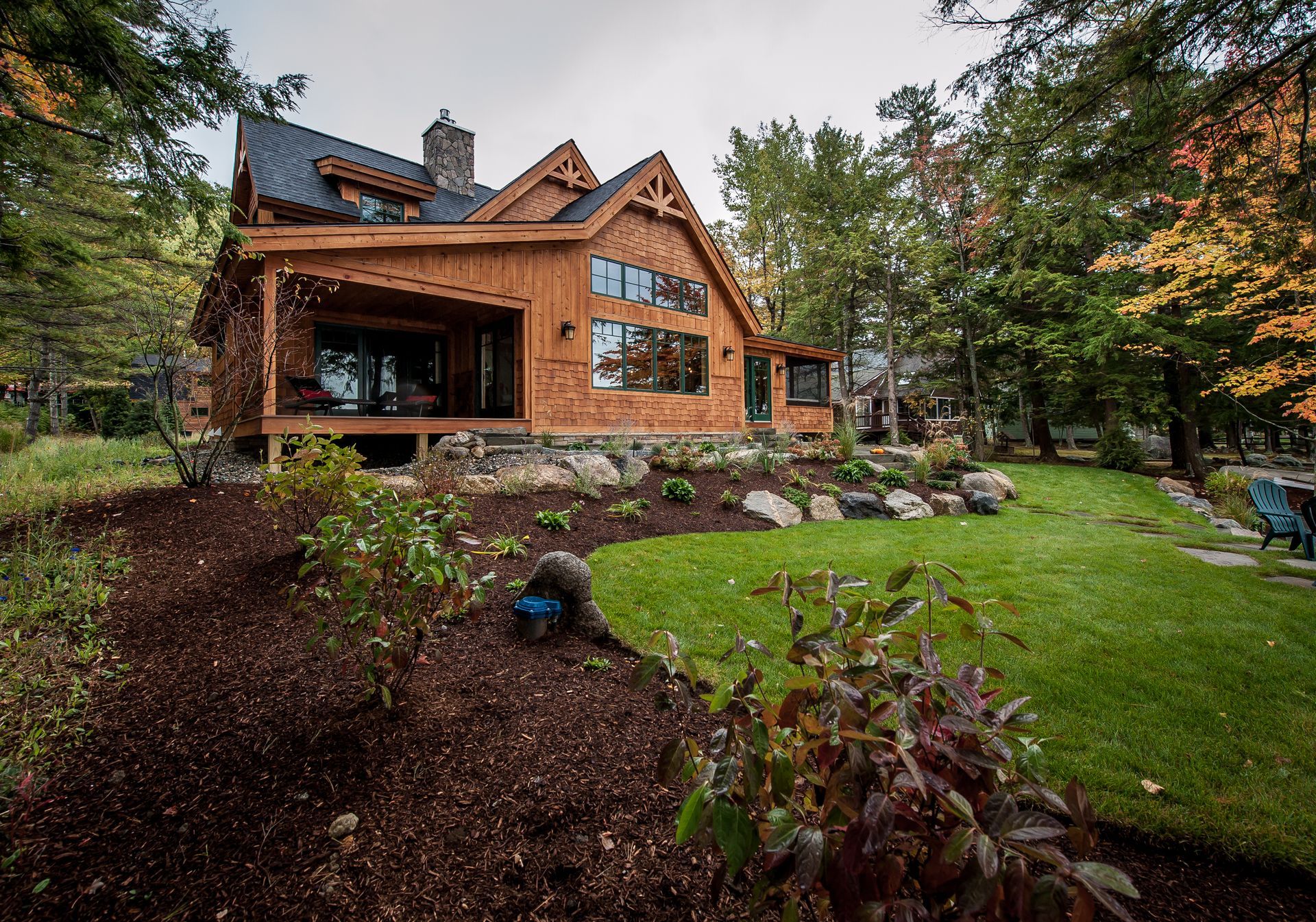 Wooden cabin with a dark roof nestled in a forest with landscaping and green grass.