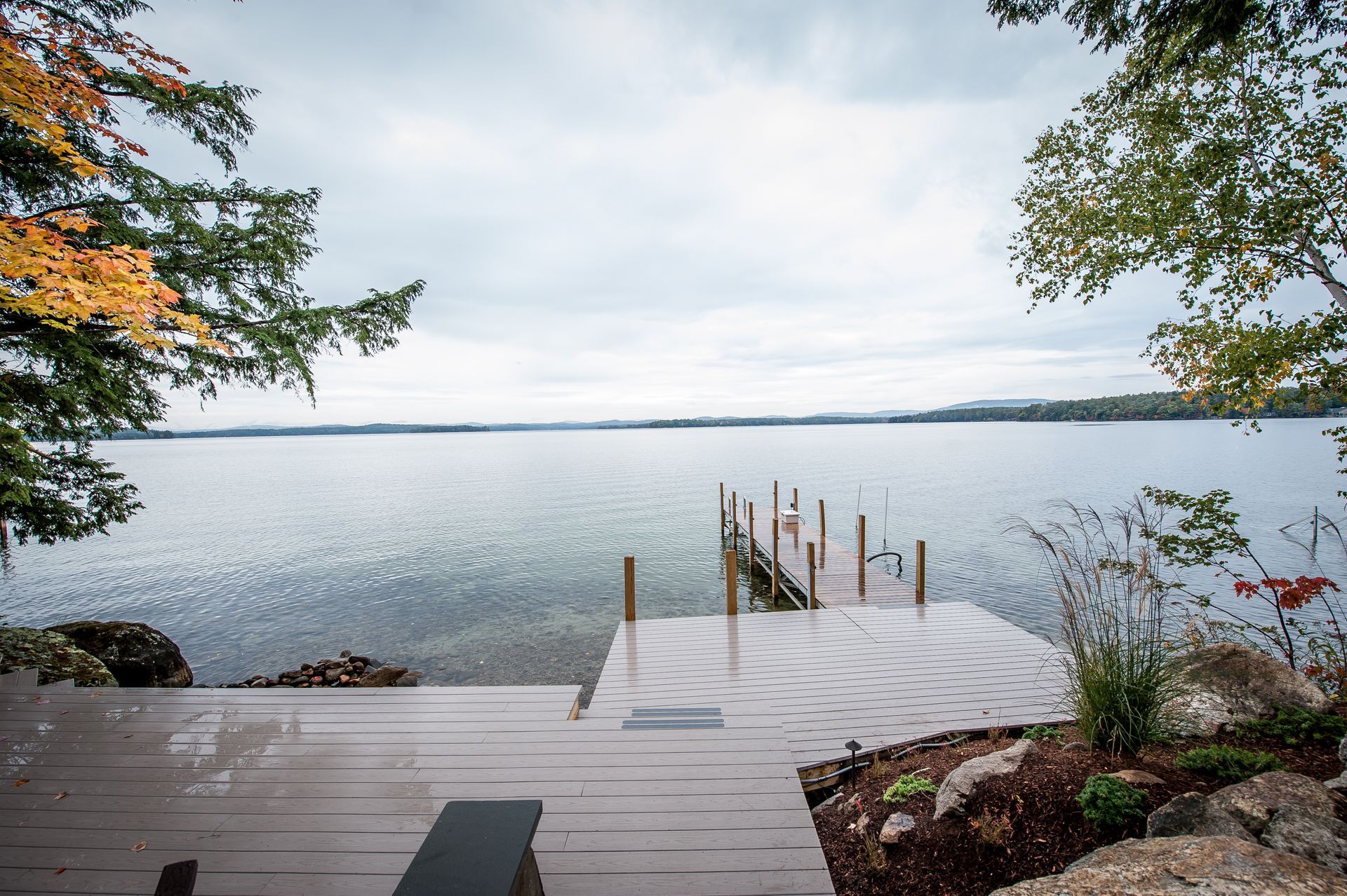 Wooden dock extending over a calm lake under a cloudy sky, autumn foliage on the edges.