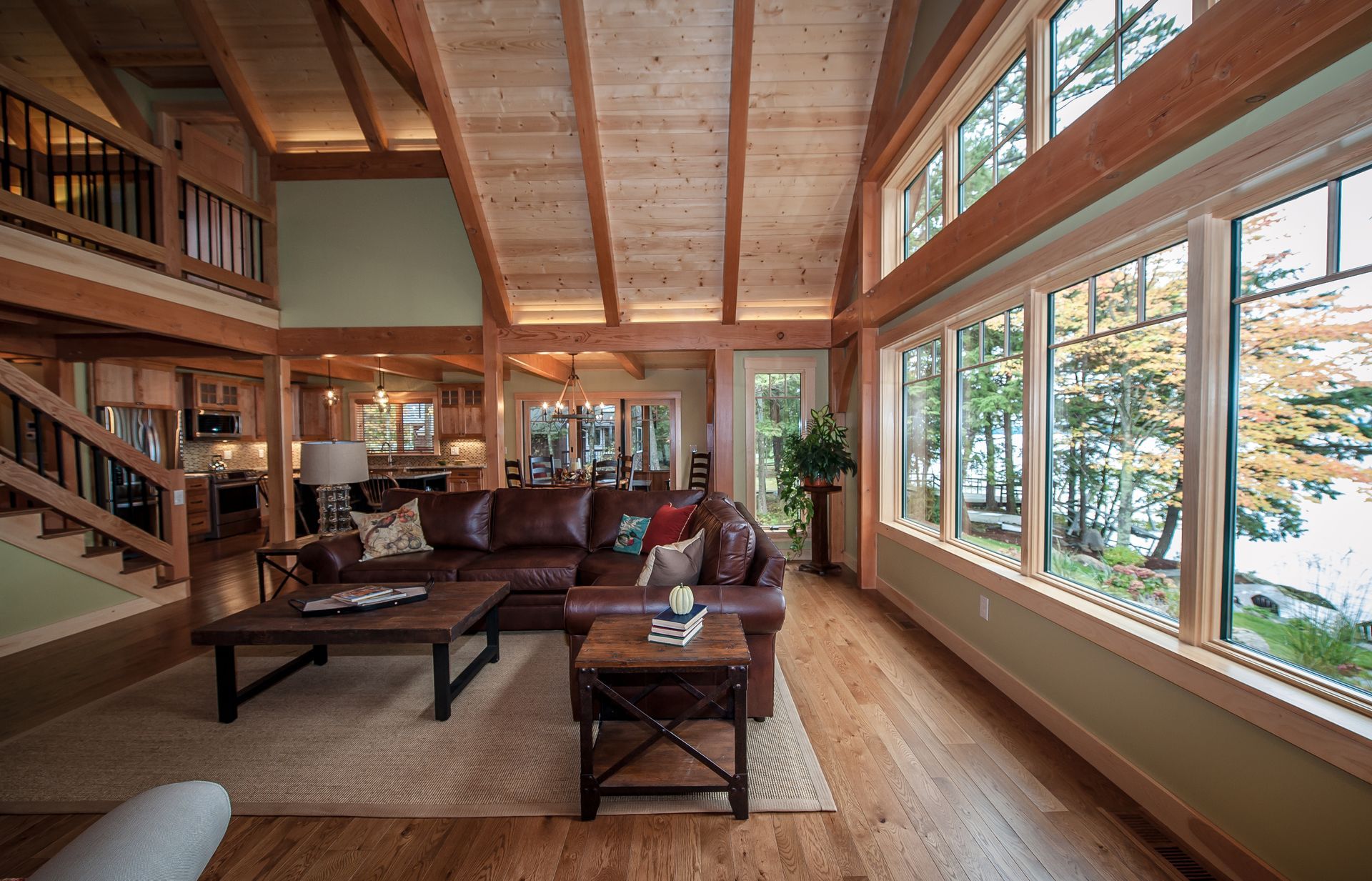 Living room with large windows, brown leather sofa, wooden beams, and a view of trees and water.