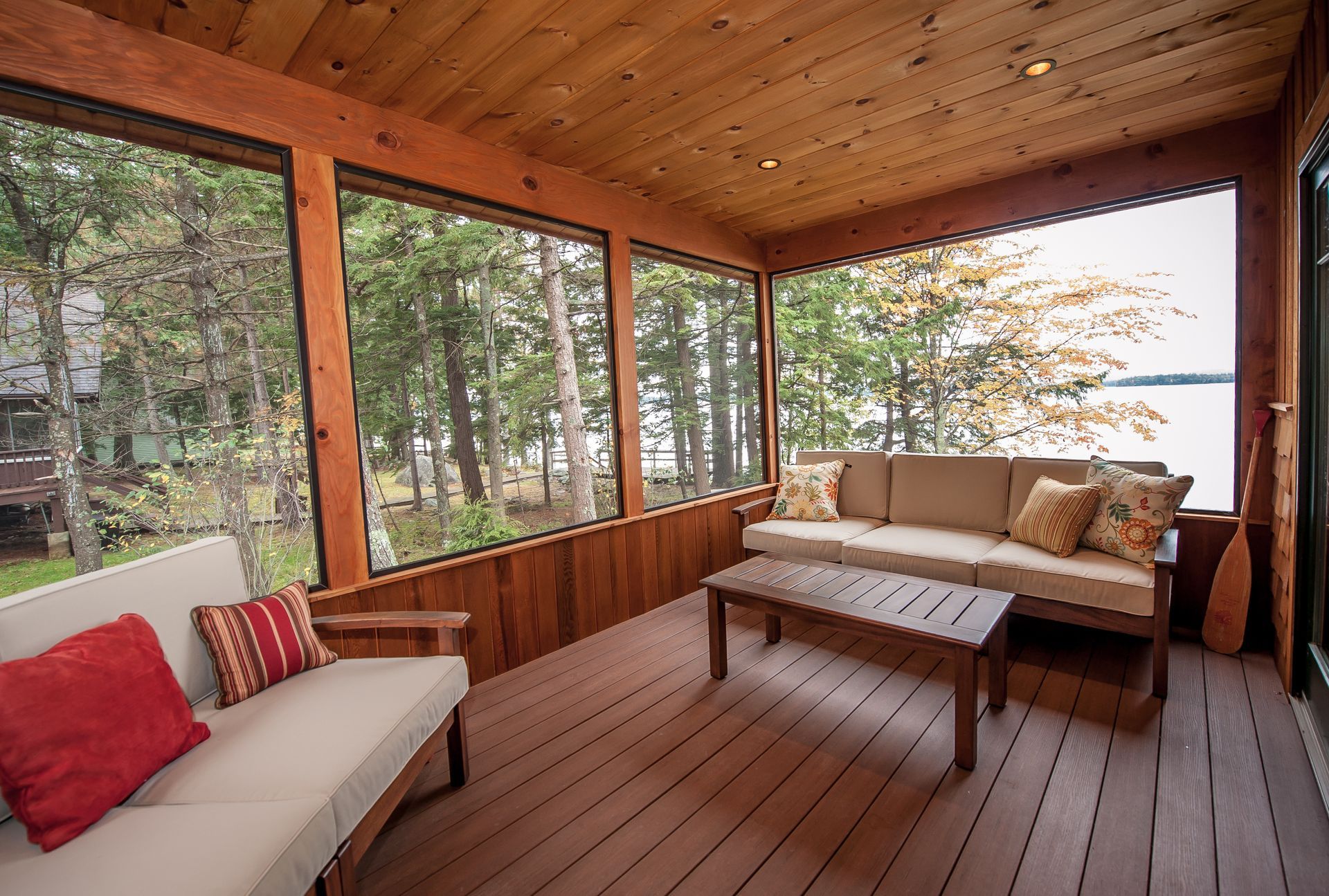 Screened-in porch with wood paneling, two beige sofas, coffee table, and view of trees and lake.