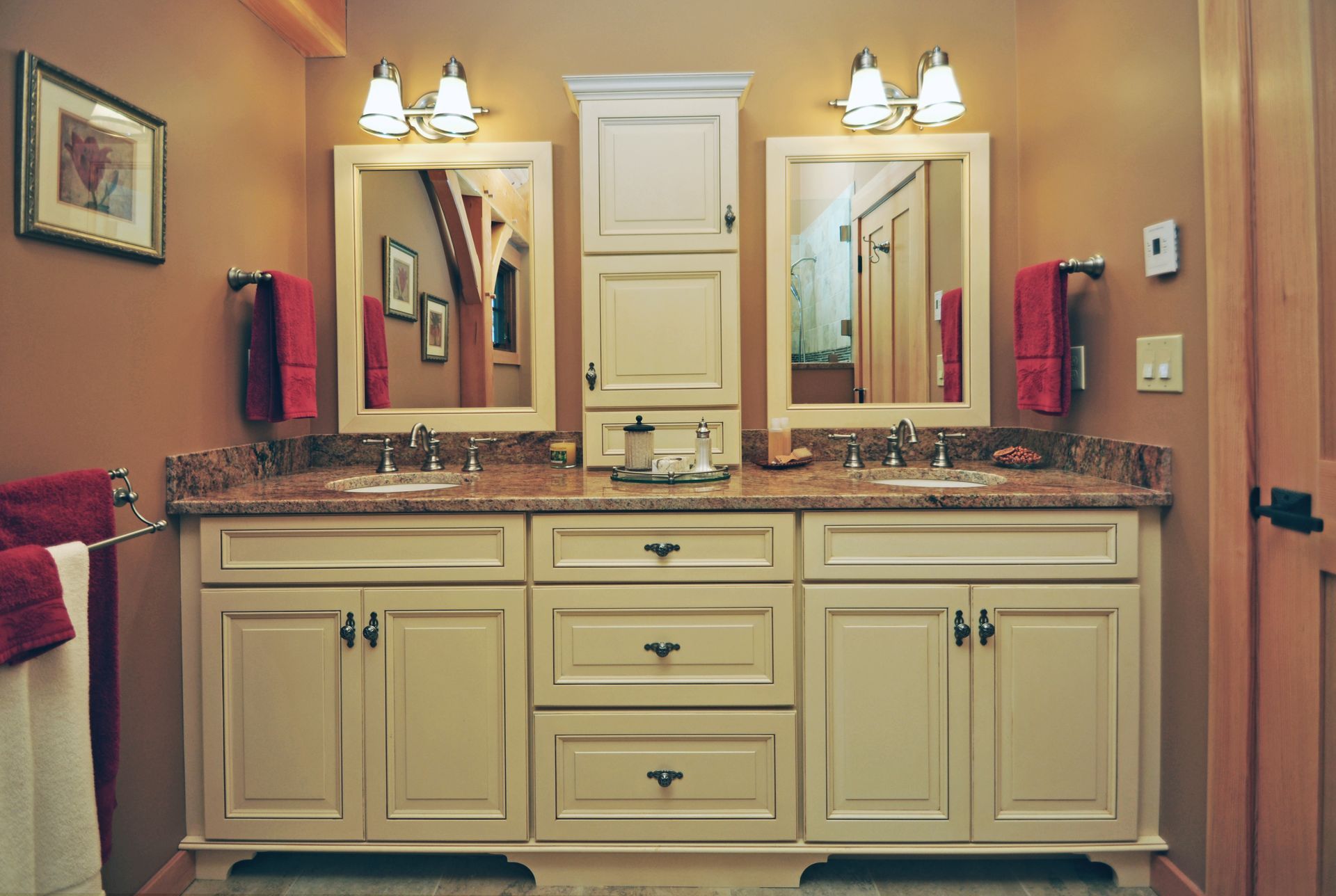 Bathroom with double sinks, light-colored cabinets, mirrors, and tan walls. Red towels and two light fixtures.