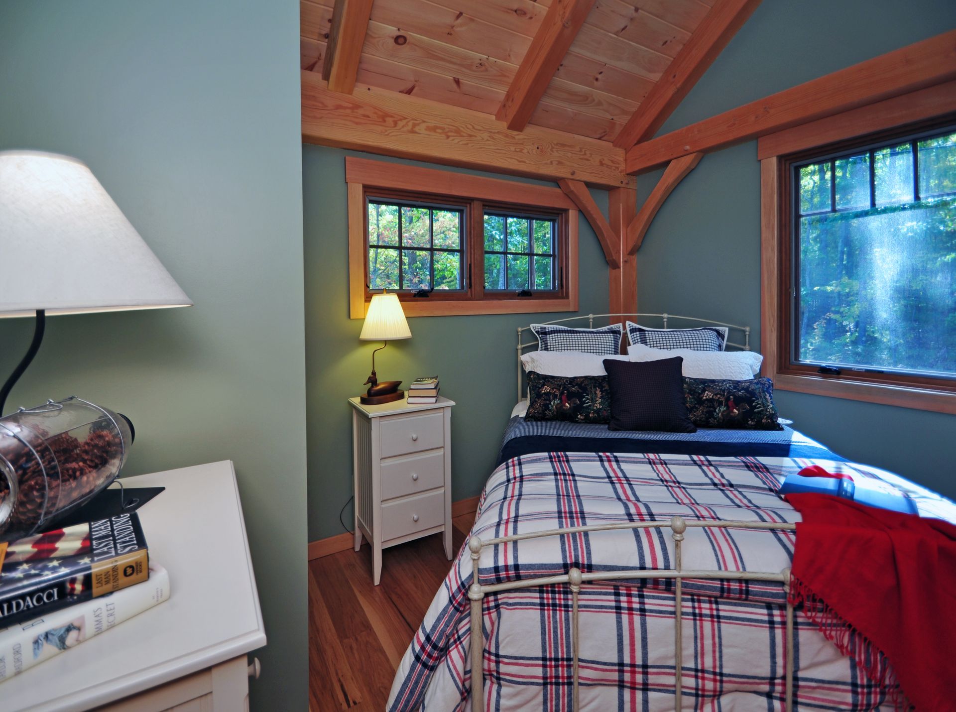 Bedroom with light blue walls, plaid bedding, wooden beams, and two windows with a forest view.