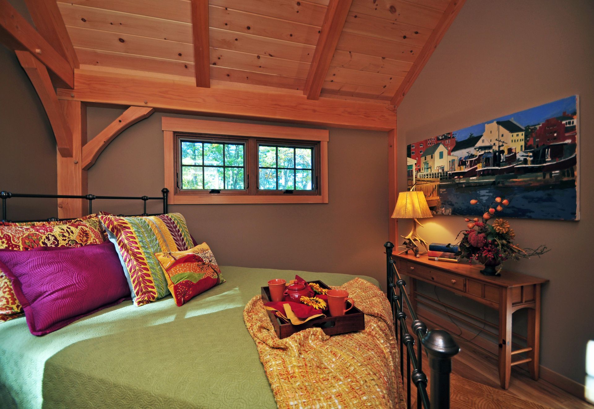 Bedroom with a green bed, colorful pillows, and a painting on the wall. Light brown wooden beams and trim.