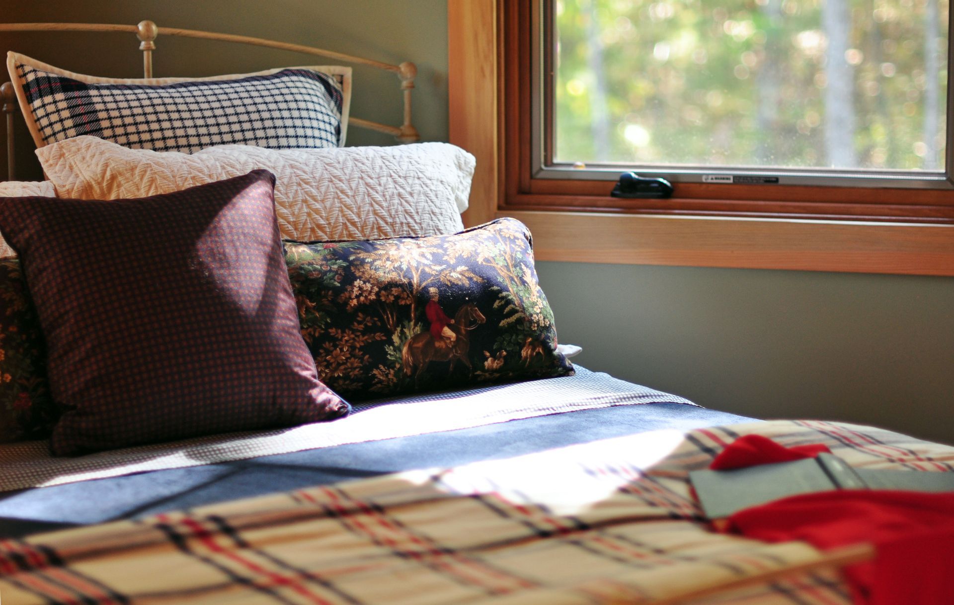 Bed with patterned pillows, plaid blanket, sunlight through window.