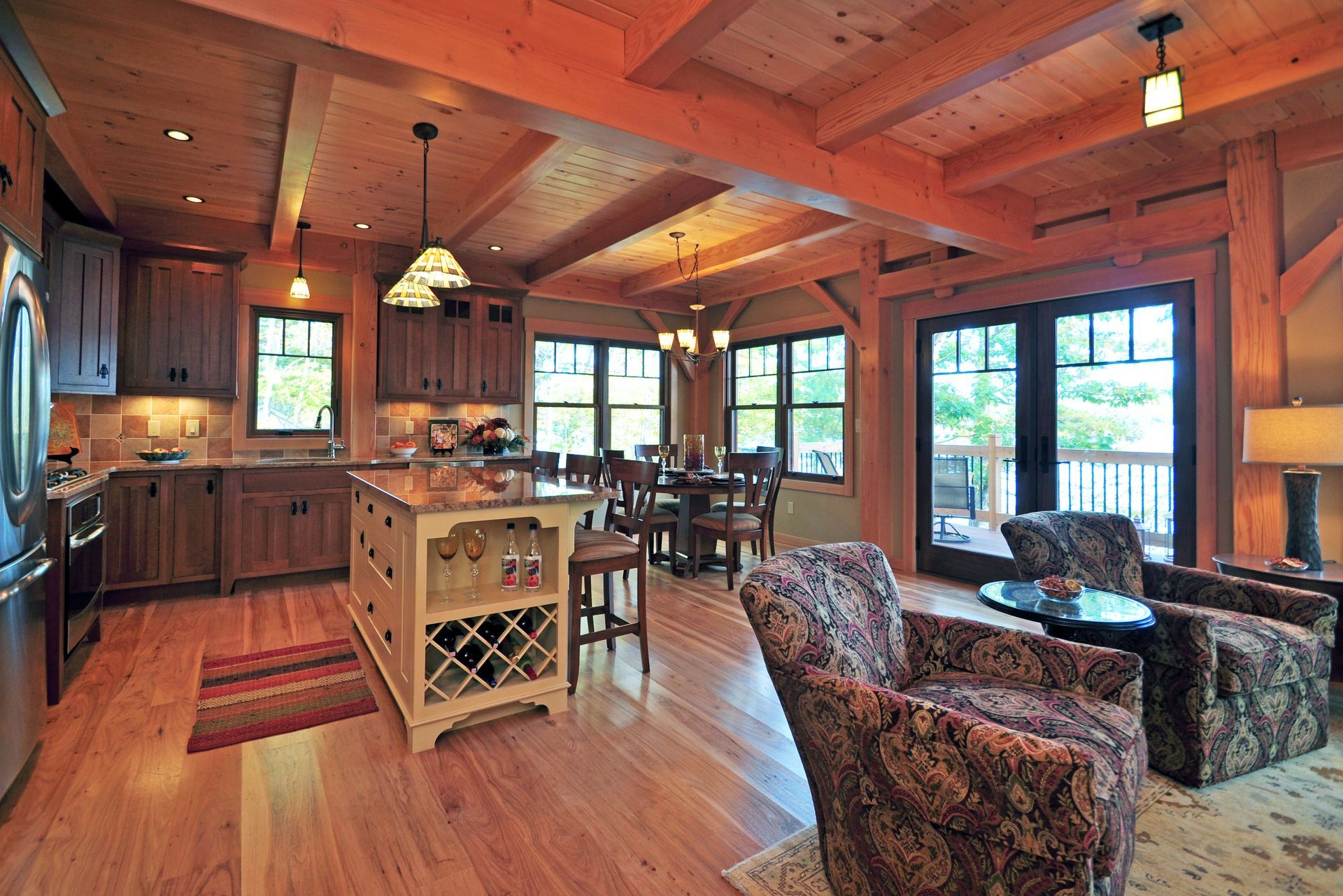 Wooden-beamed interior with kitchen, dining area, and living room. Brown cabinets, island, and wood floors. Two patterned armchairs.