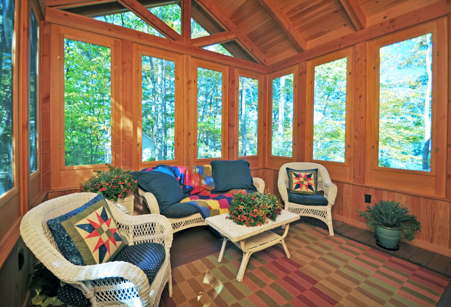 Sunroom with wicker furniture, wood paneling, and large windows overlooking trees.
