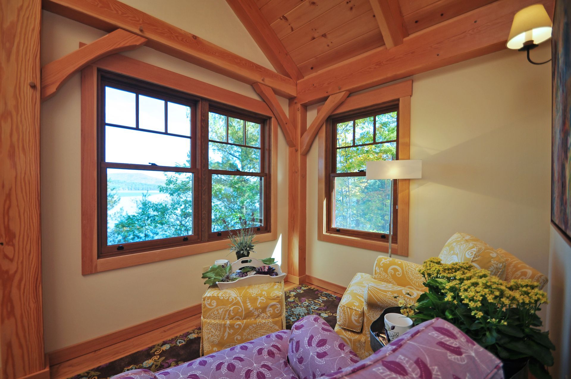 Cozy corner with wood beams and two windows overlooking a lake. Yellow armchair, purple floral couch, and flowers.