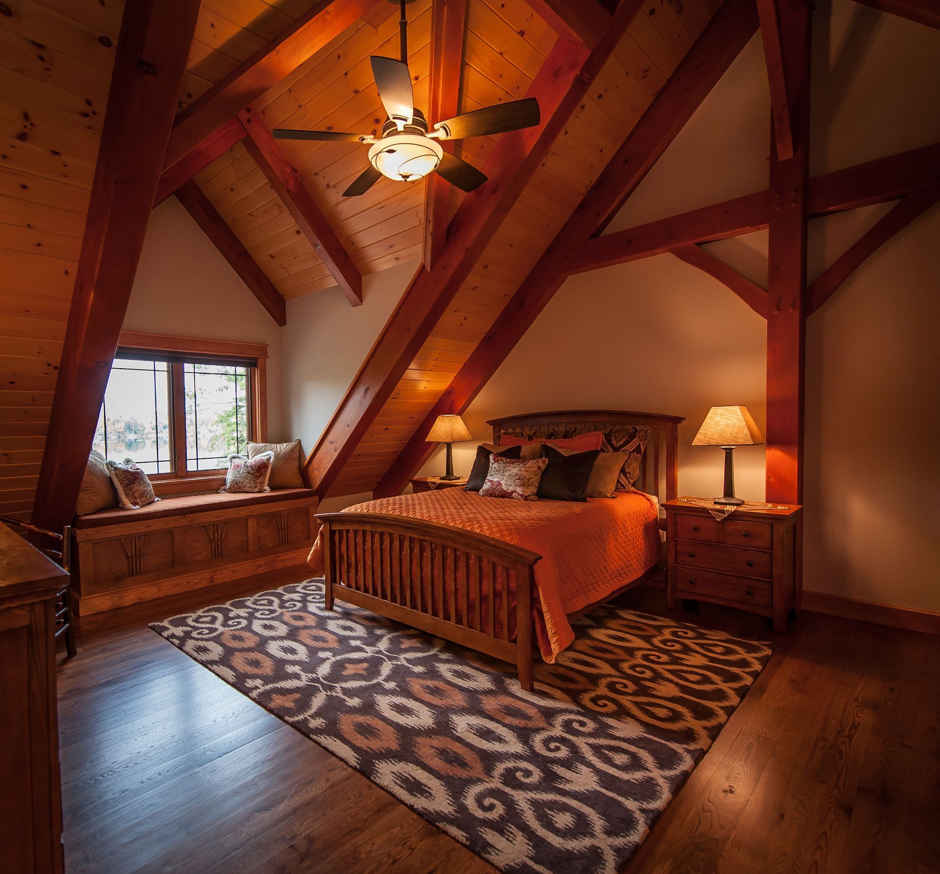 Bedroom with wooden beams, bed, rug, window seat, and lamps.