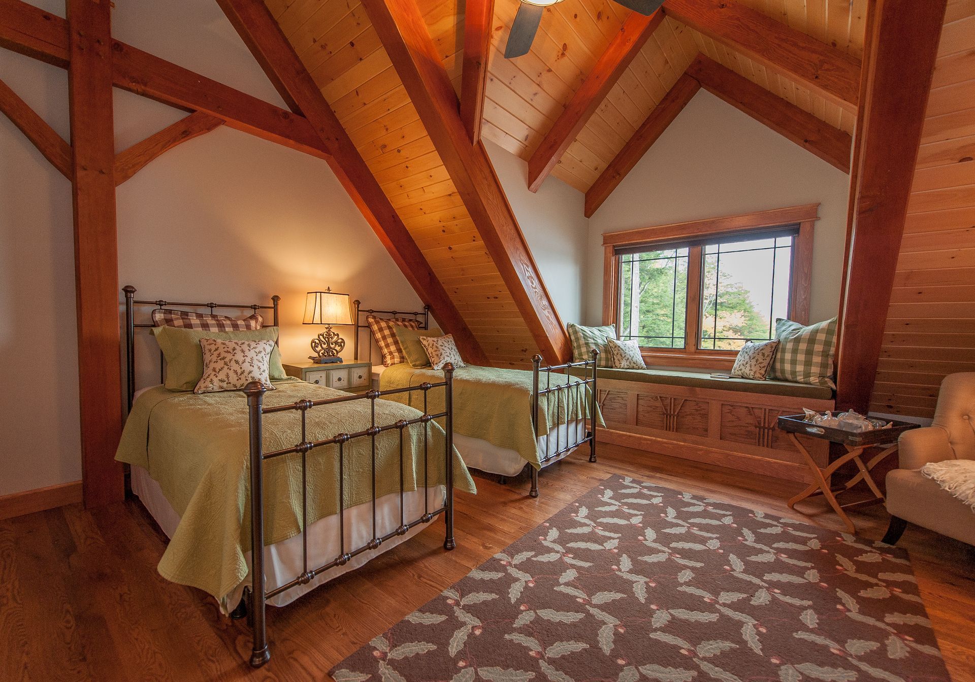 Bedroom with two twin beds under a sloped ceiling, wooden beams, window seat, and patterned rug.