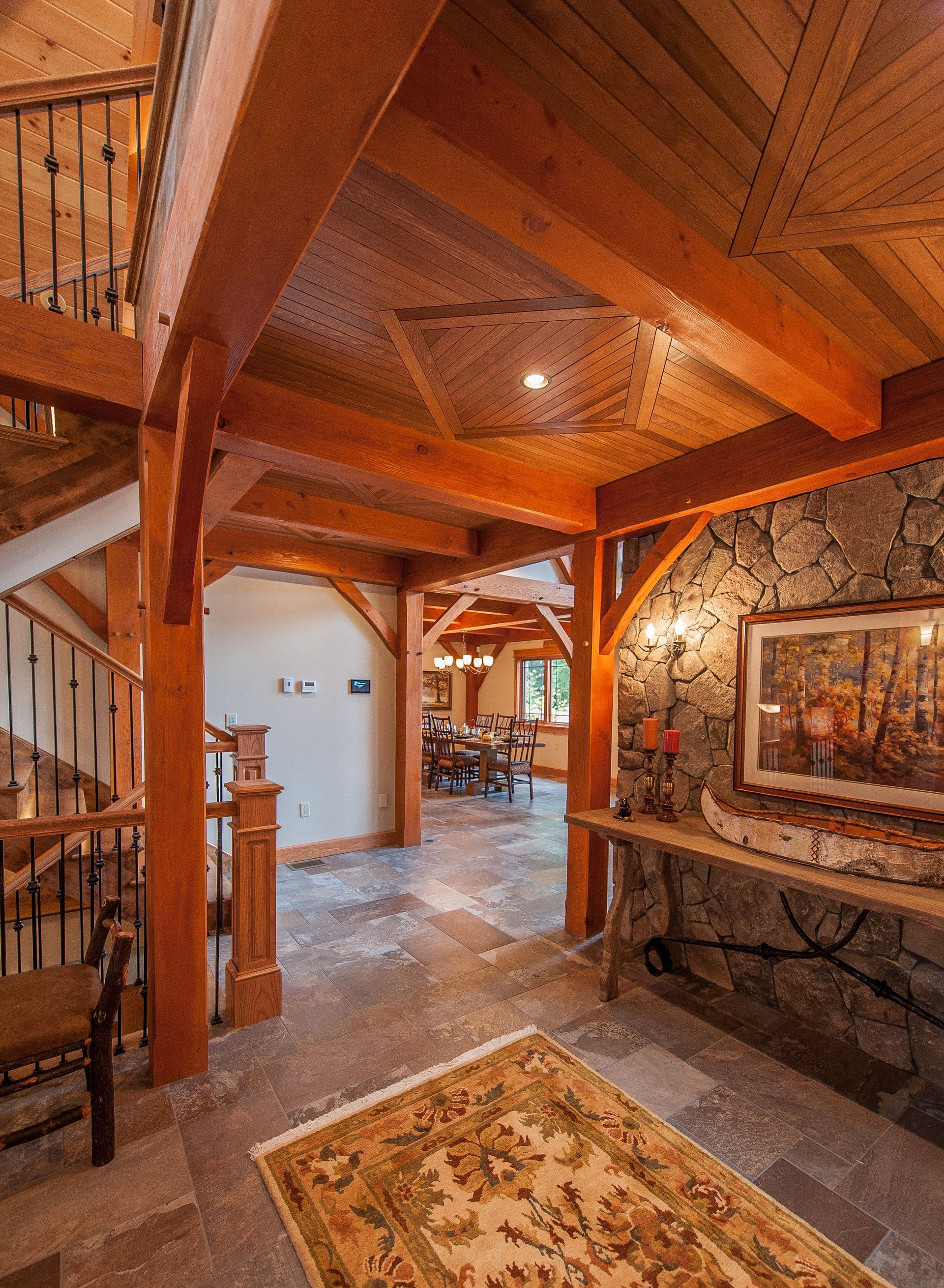 Rustic foyer with exposed wood beams, stone wall, and ornate rug. Staircase on the left.