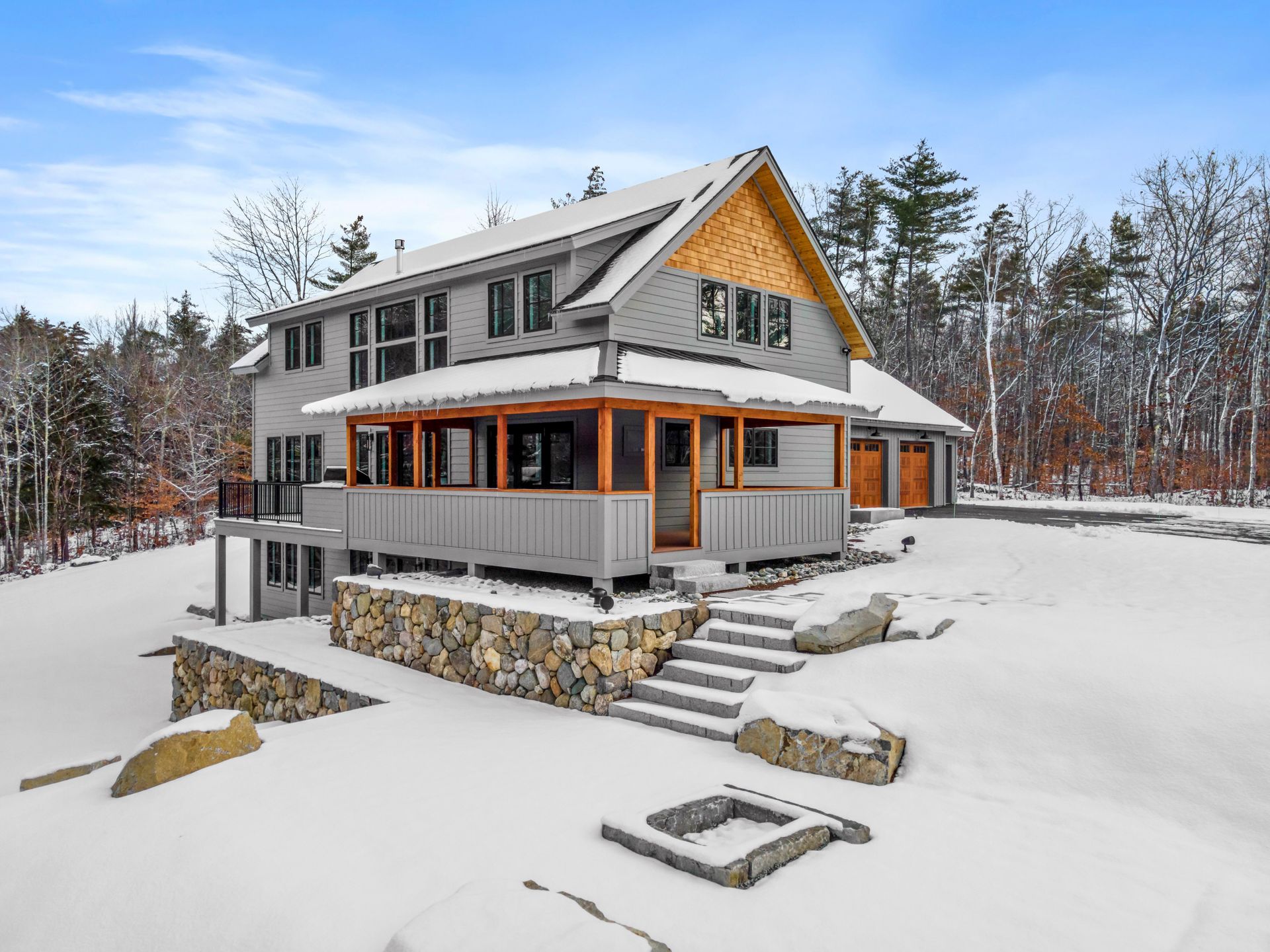 Two-story gray house with wood accents, porch, and stone foundation, set in a snowy landscape.