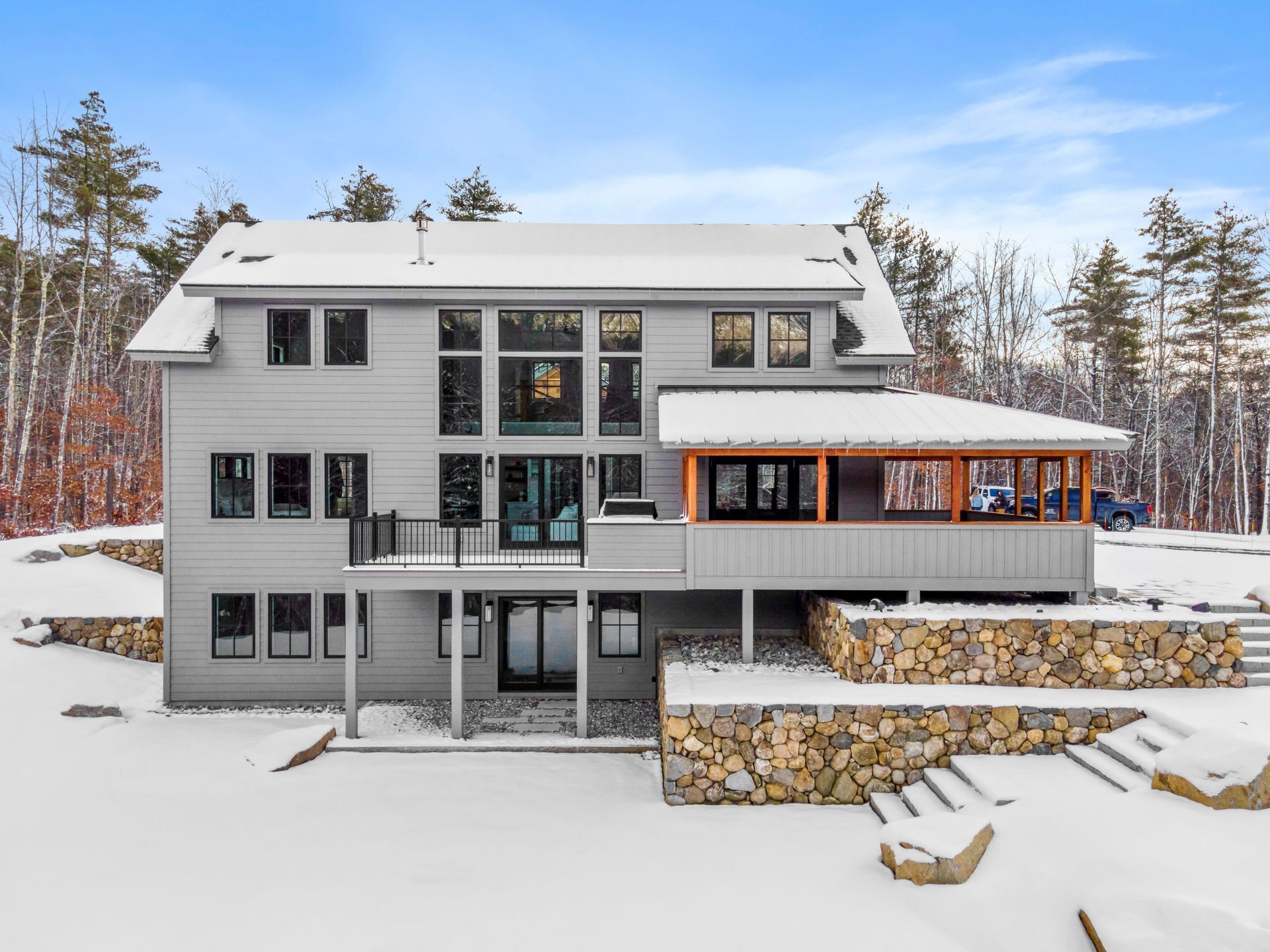 Two-story gray house with large windows, deck, and covered porch; set in snowy landscape.