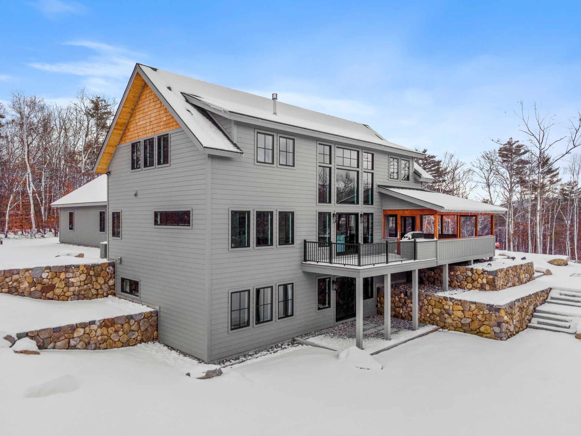 Two-story gray house with a snow-covered yard and stone retaining walls in a winter setting.