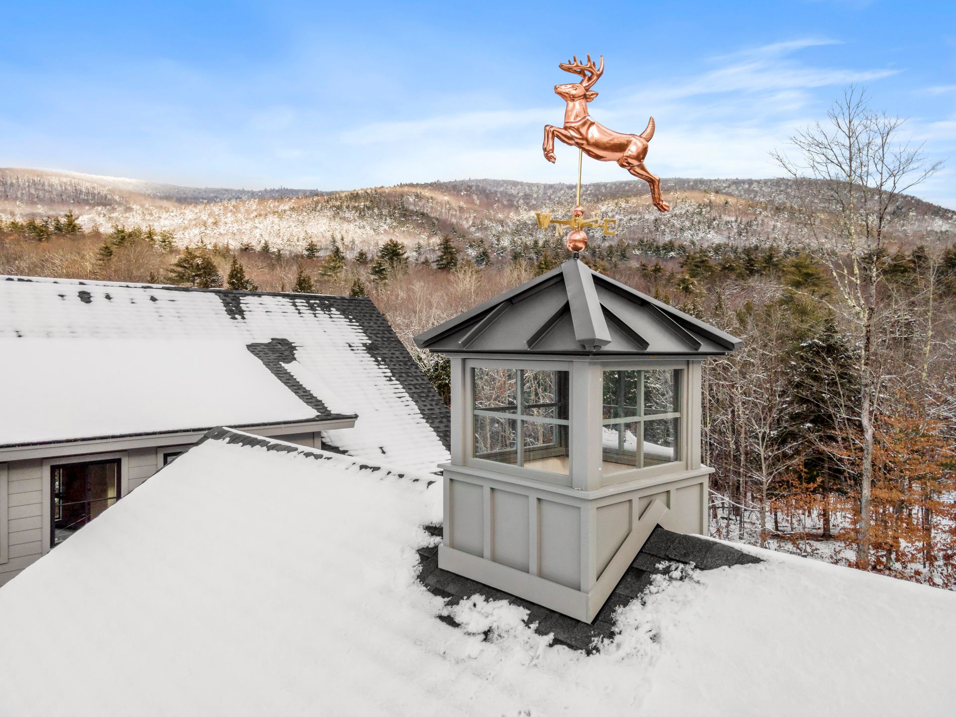 Snowy rooftop with copper deer weathervane atop a cupola, forested hills in the background.