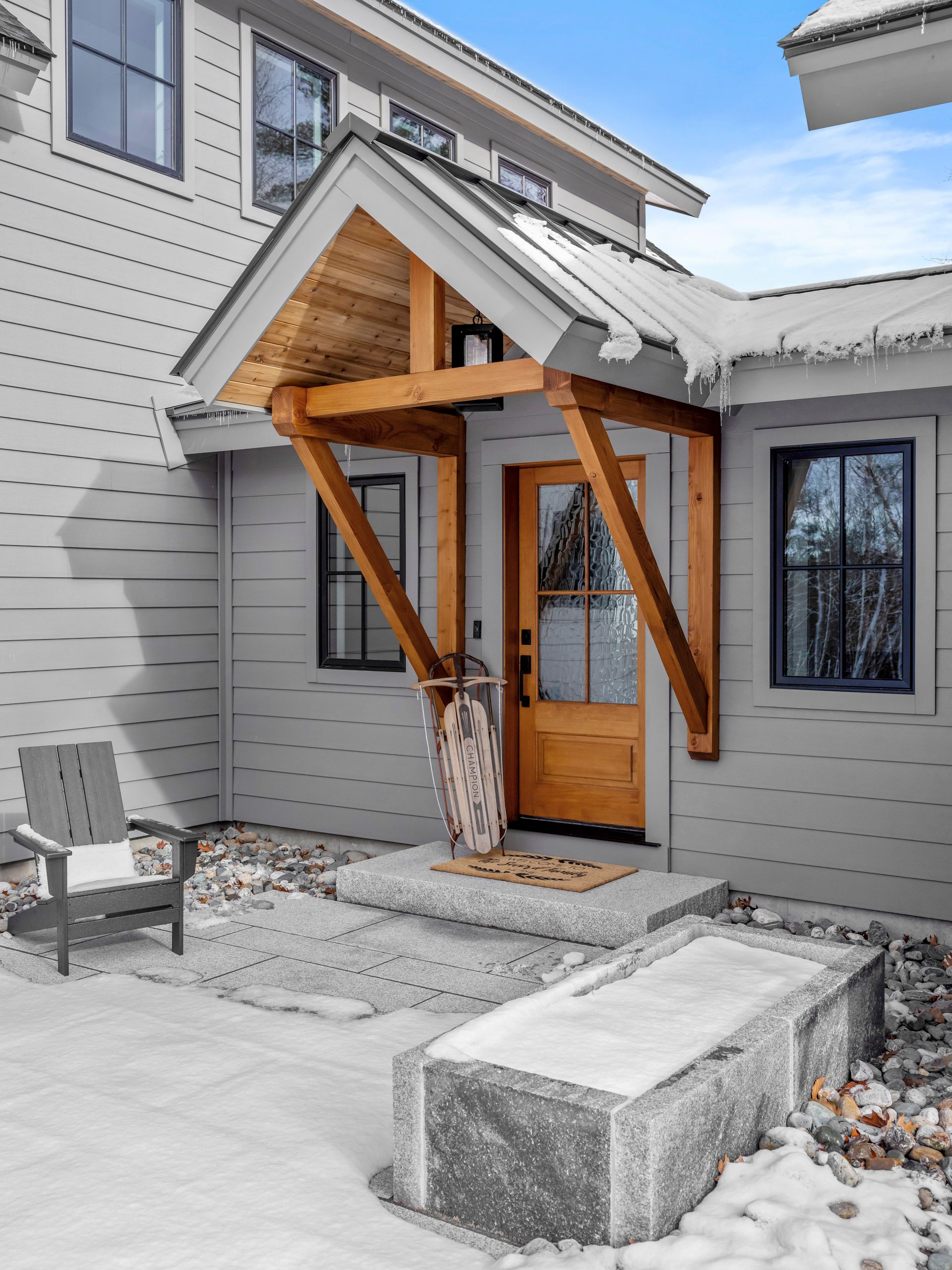 Gray house entrance with wooden door, porch, and snow-covered stone bench.