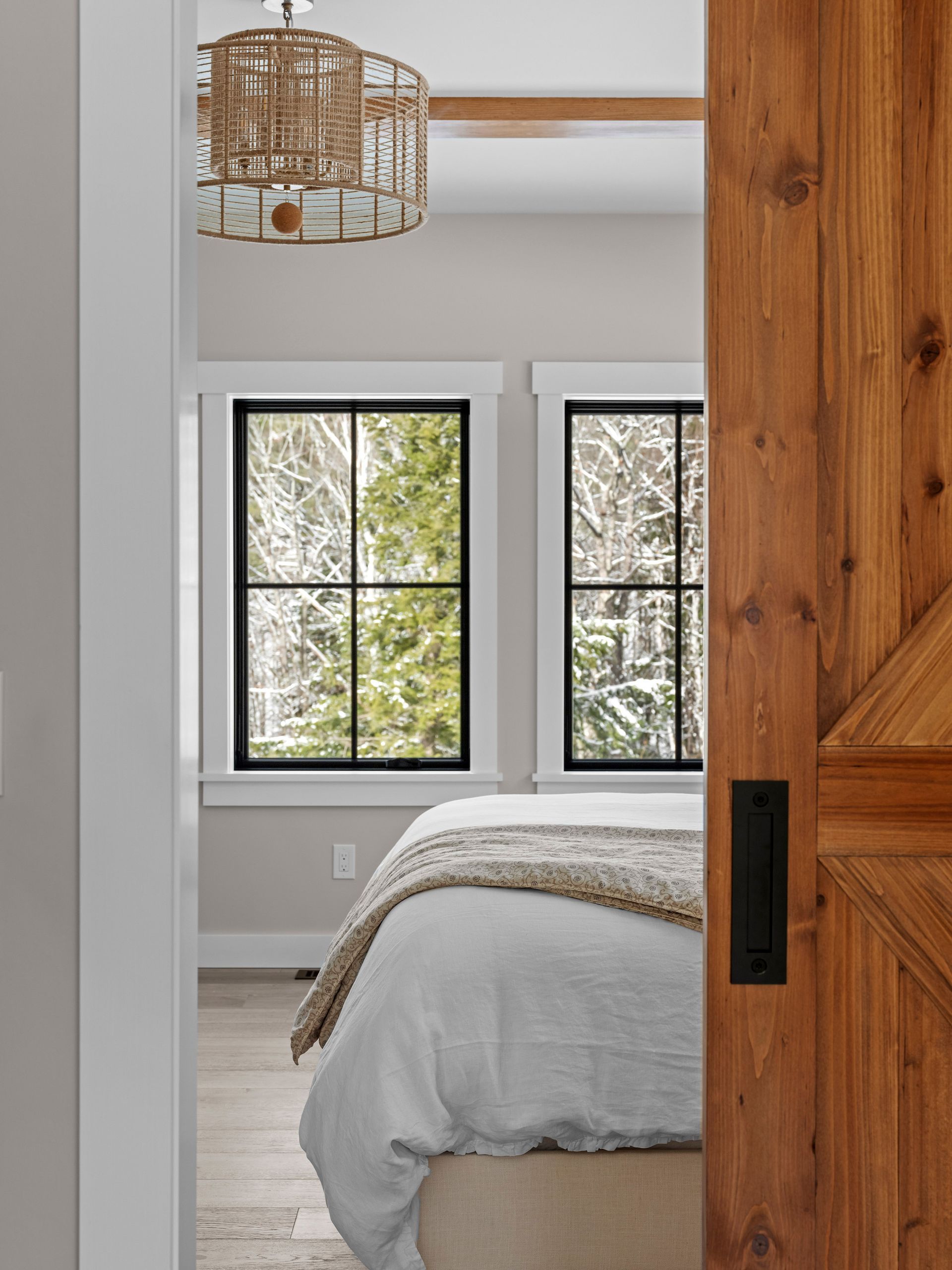 Bedroom interior with a partially open wooden door, two windows with a view of trees, and a bed.