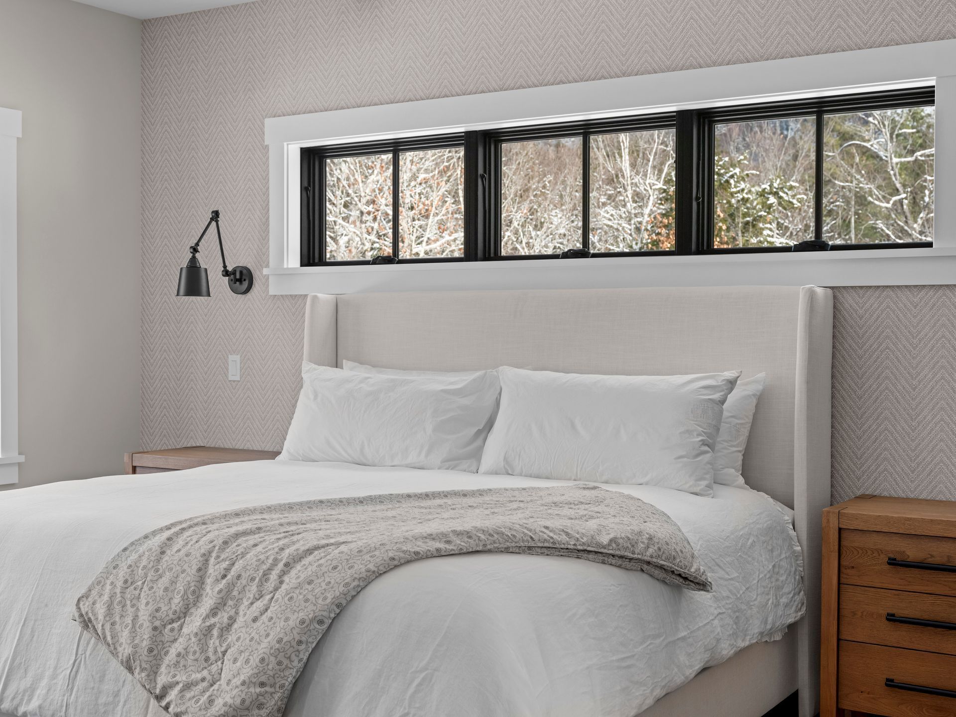 Bedroom with white bed, gray throw, black-framed windows, and a wall-mounted lamp.