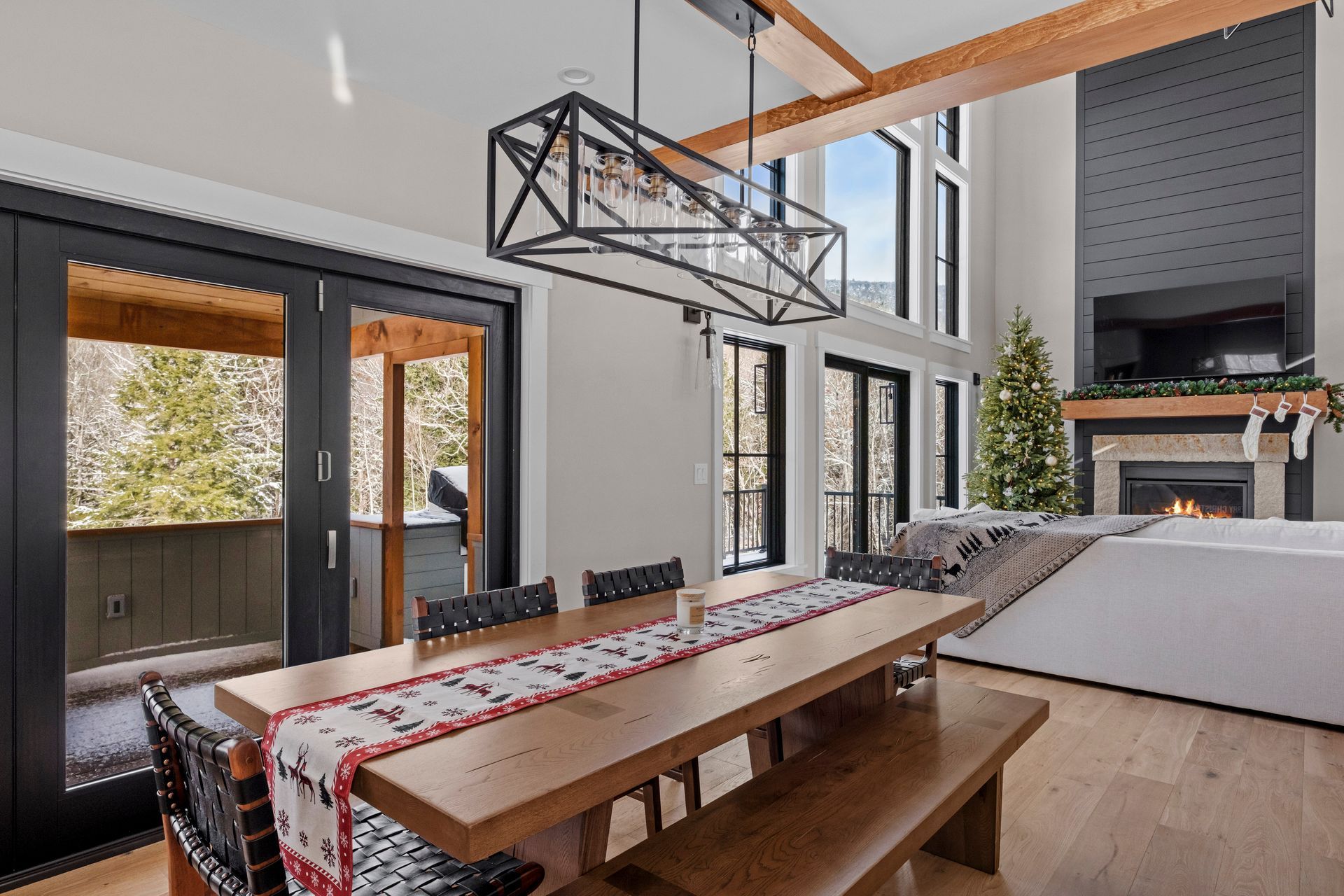 Dining area with large table, bench, chairs, and fireplace, near glass doors and a Christmas tree.