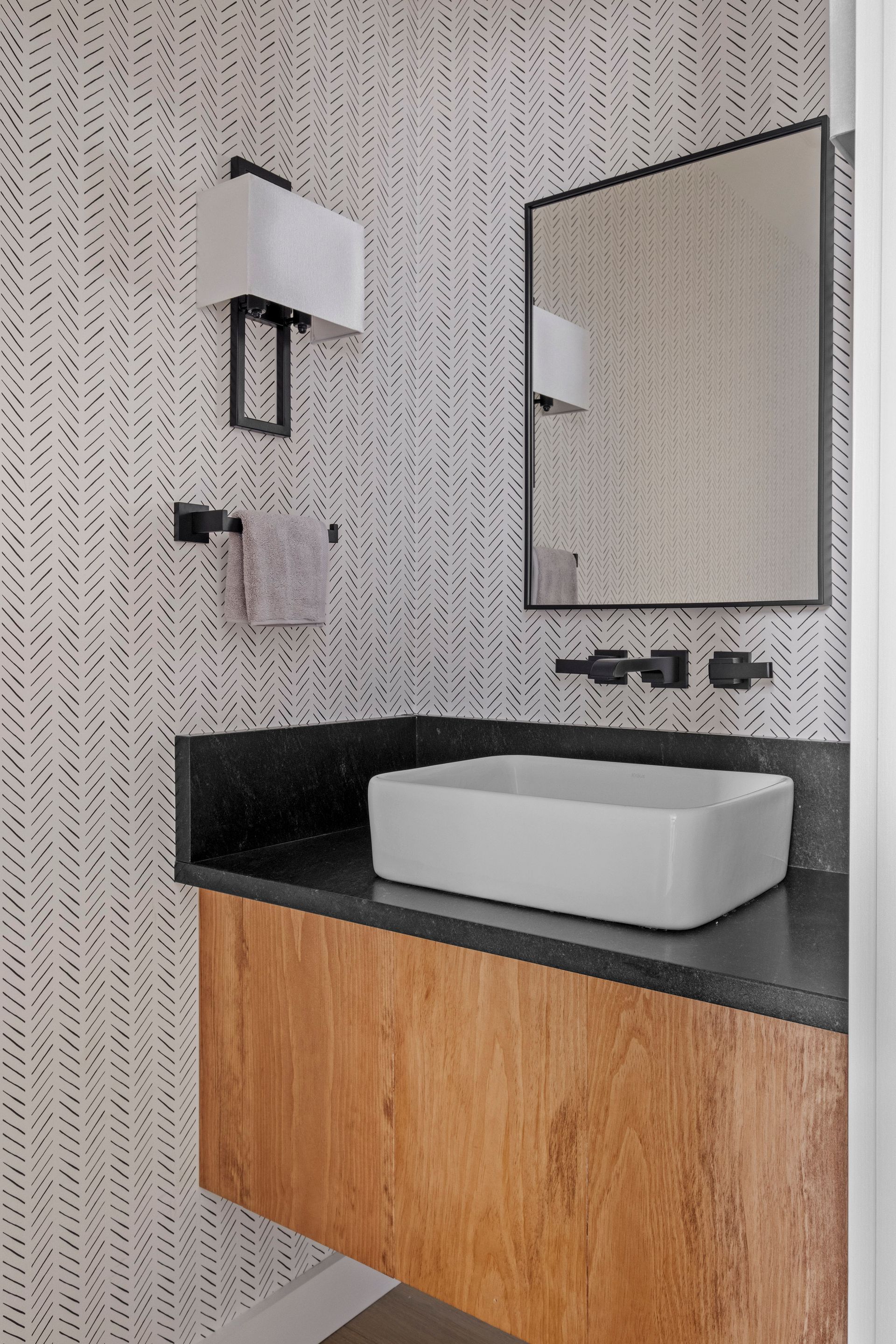 Modern bathroom with wood vanity, rectangular sink, black countertop, patterned wallpaper, and black-framed mirror.