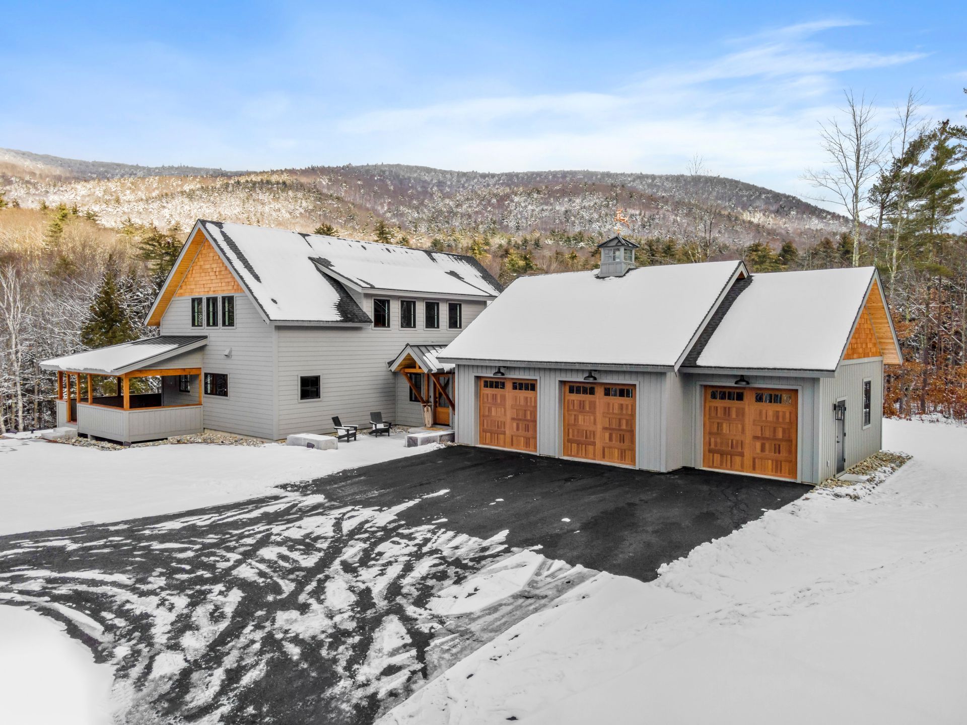 Snow-covered house and detached garage with brown doors, black asphalt driveway, mountain backdrop. Winter scene.