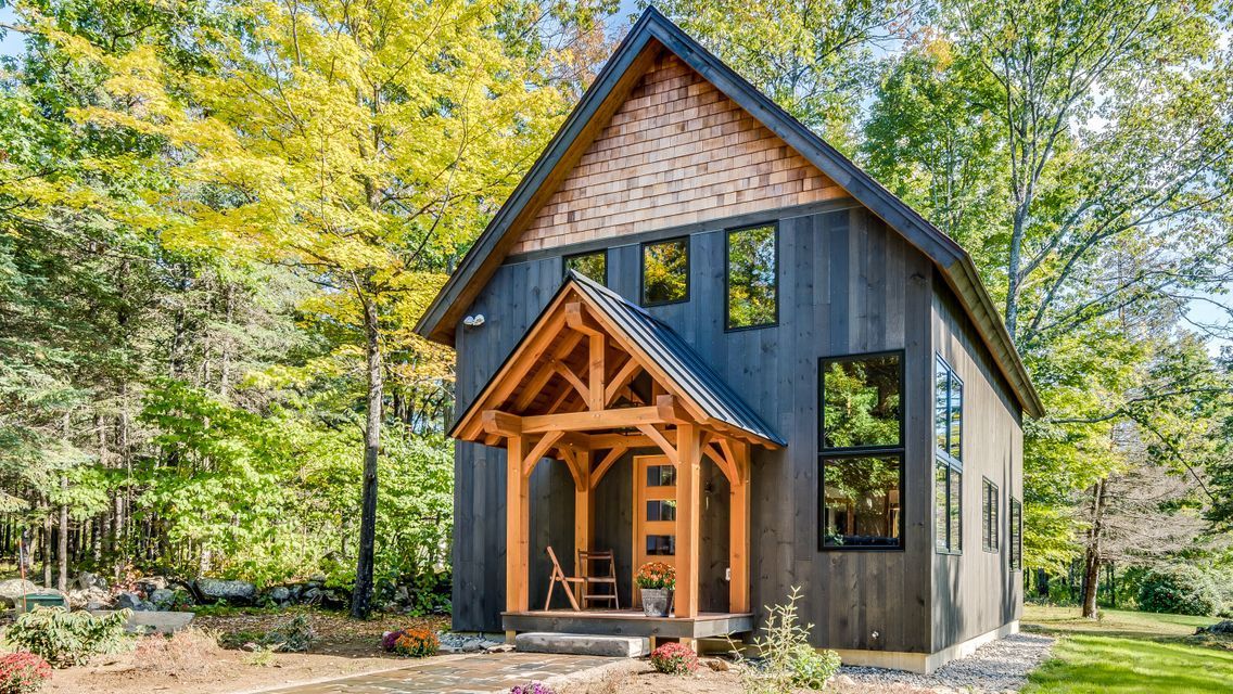 A small black house with a wooden porch in the middle of a forest.