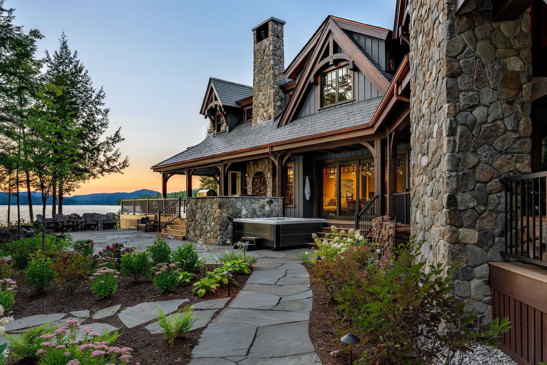 A luxury stone house with a metal roof and hot tub overlooks a lake at sunset, featuring a stone patio and landscaping.