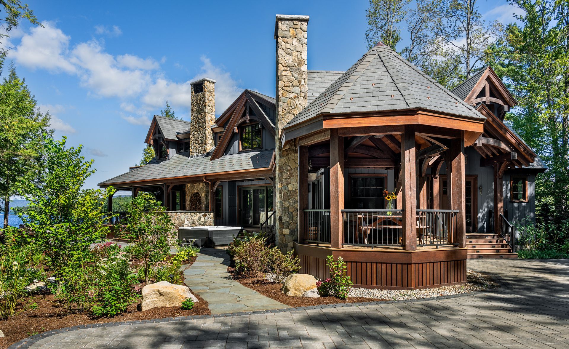 A rustic, stone-and-timber luxury home featuring a covered hexagonal porch, lush landscaping, and a scenic lake view.