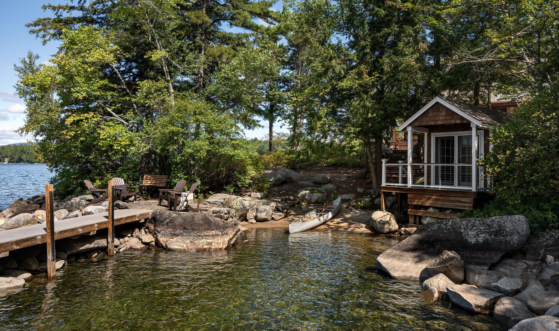 A rustic wooden cabin sits on a rocky shoreline by a lake, next to a wooden dock and surrounded by lush green trees.