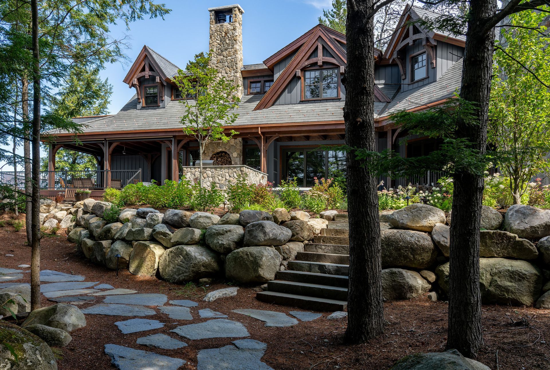A rustic, multi-gabled house with gray siding and a stone chimney, set behind a large boulder retaining wall and trees.