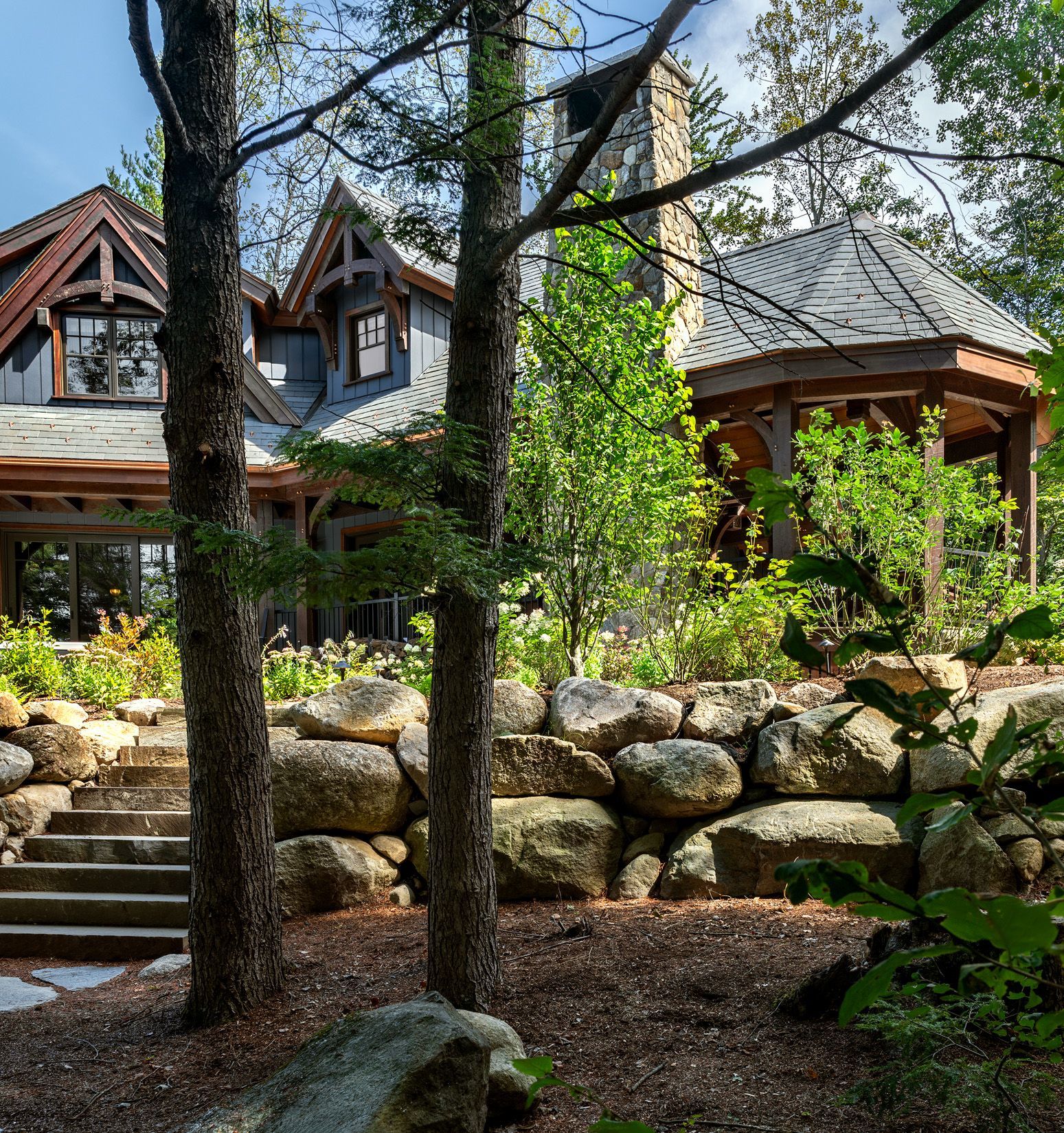A large house with stone siding and a rustic gazebo sits behind a stone retaining wall and staircase in a wooded area.